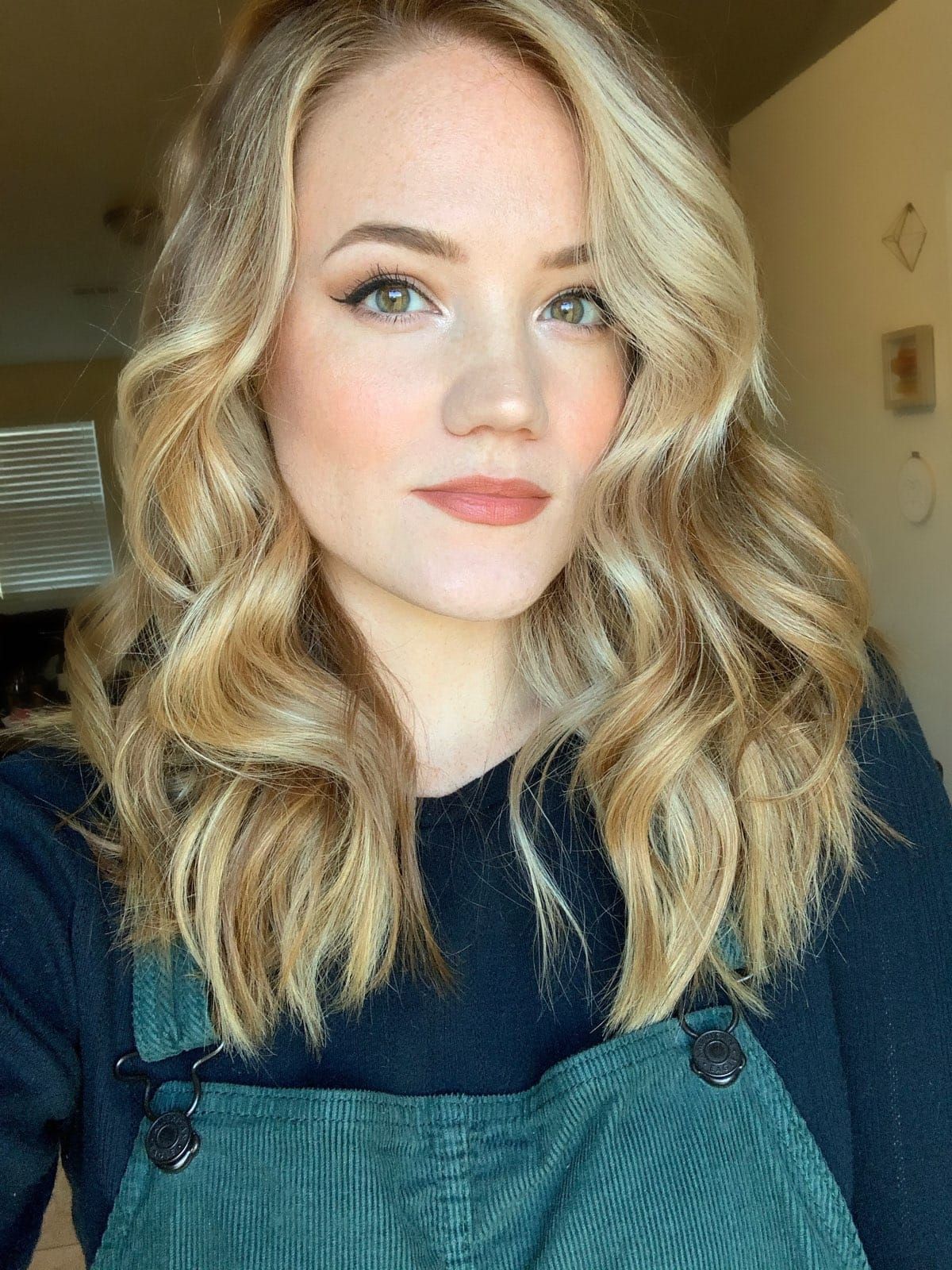 A person with shoulder-length, blonde wavy hair, wearing a dark shirt under green overalls, posing for a selfie.