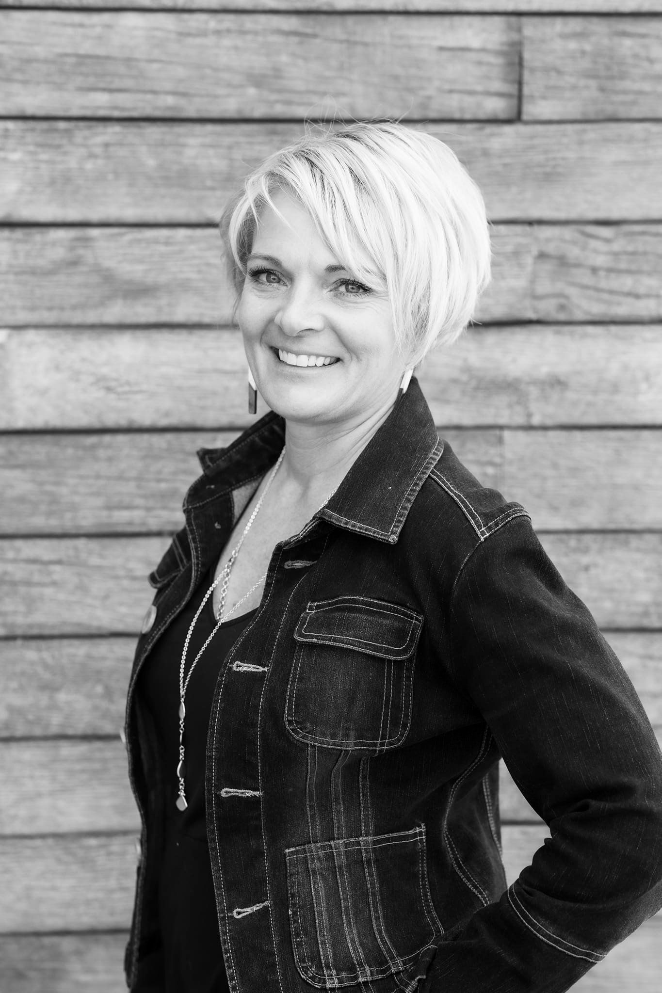 A smiling person with a short, layered haircut poses against a horizontal wood-plank wall while wearing a dark jacket.