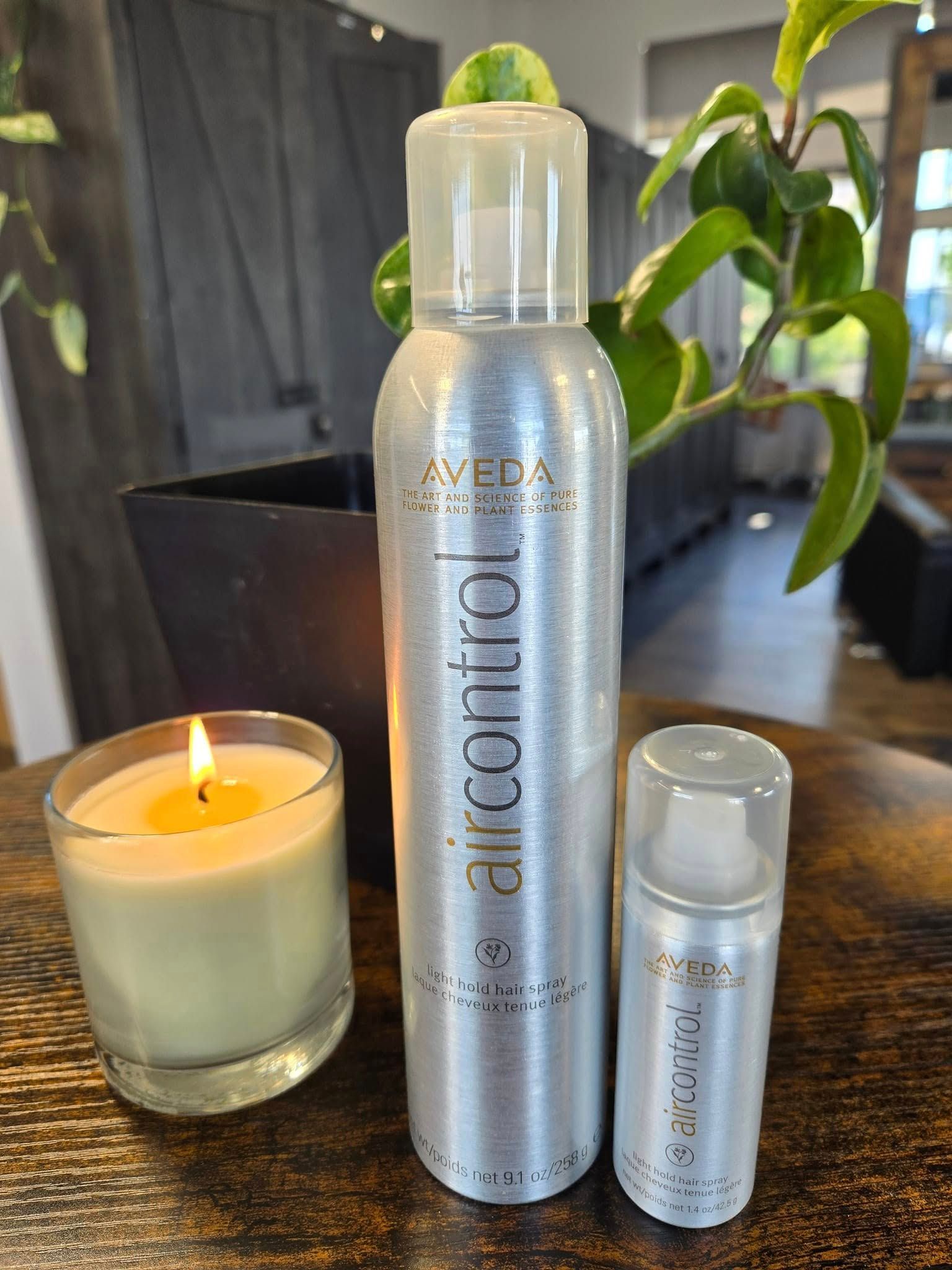 A tall bottle of Aveda Air Control hairspray, a matching travel-sized bottle, and a lit candle sit on a wooden table.