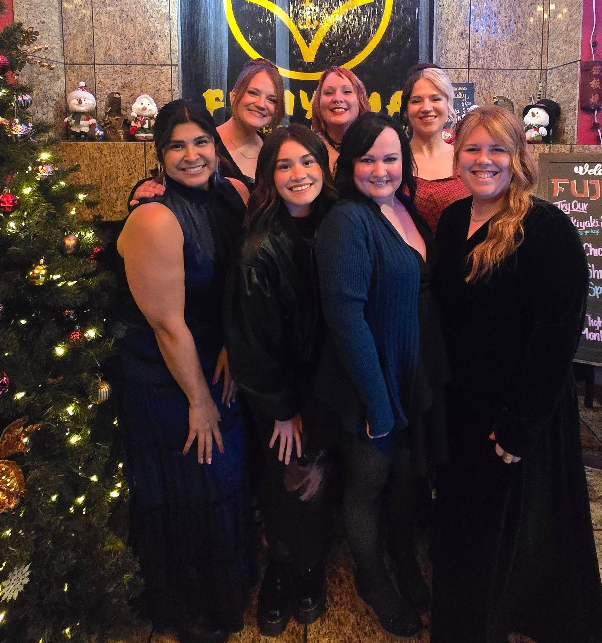 Six people stand smiling together in front of a decorated Christmas tree and a sign in an indoor setting.