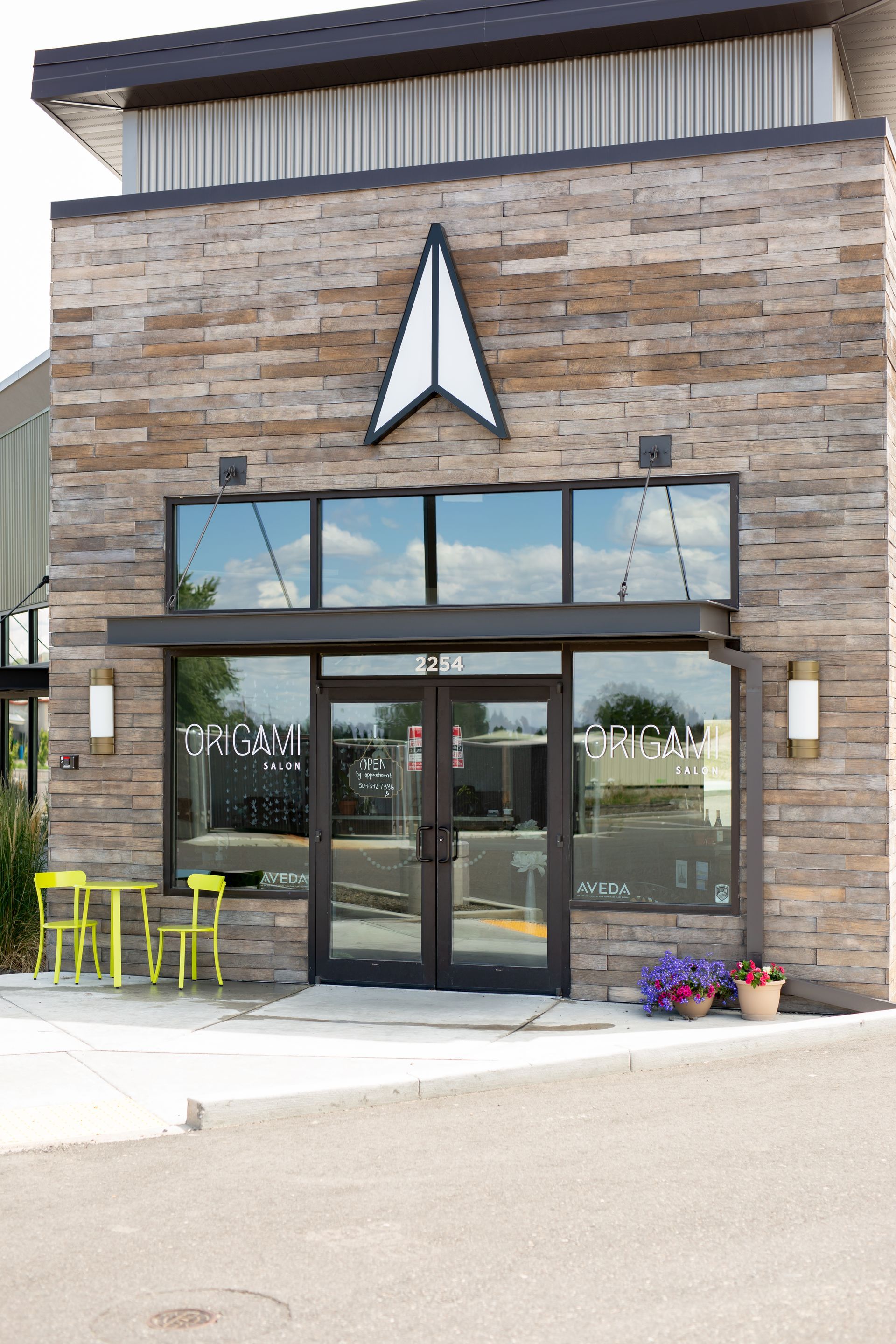 The entrance of a stone building with a prominent triangular logo, glass doors, and yellow chairs on the patio.