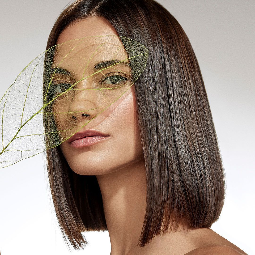 A person with crimped brown hair looks at the camera through a transparent, veined green leaf.