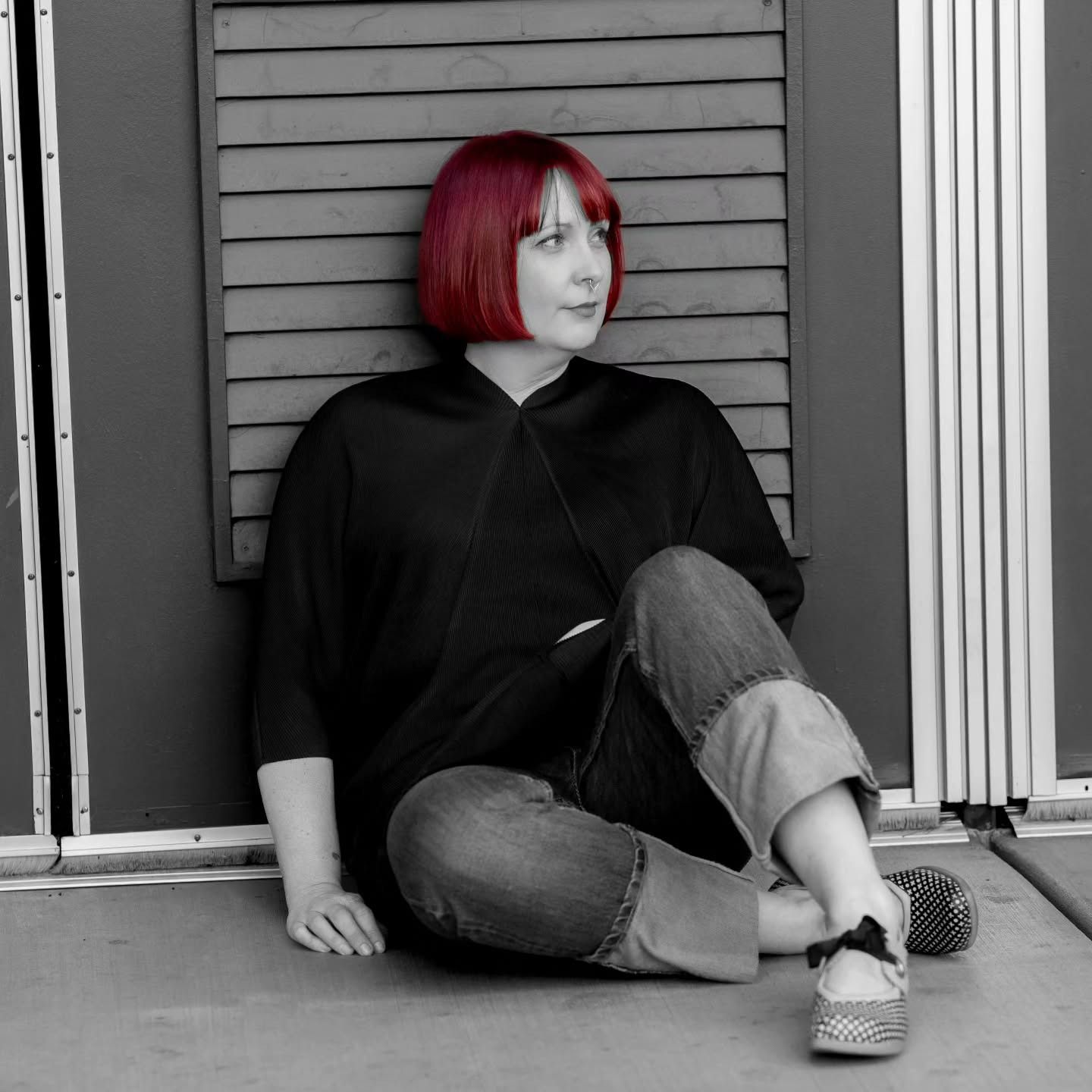A person with a red bob haircut sits on the floor against a slatted backdrop, wearing a black top and cuffed blue jeans.