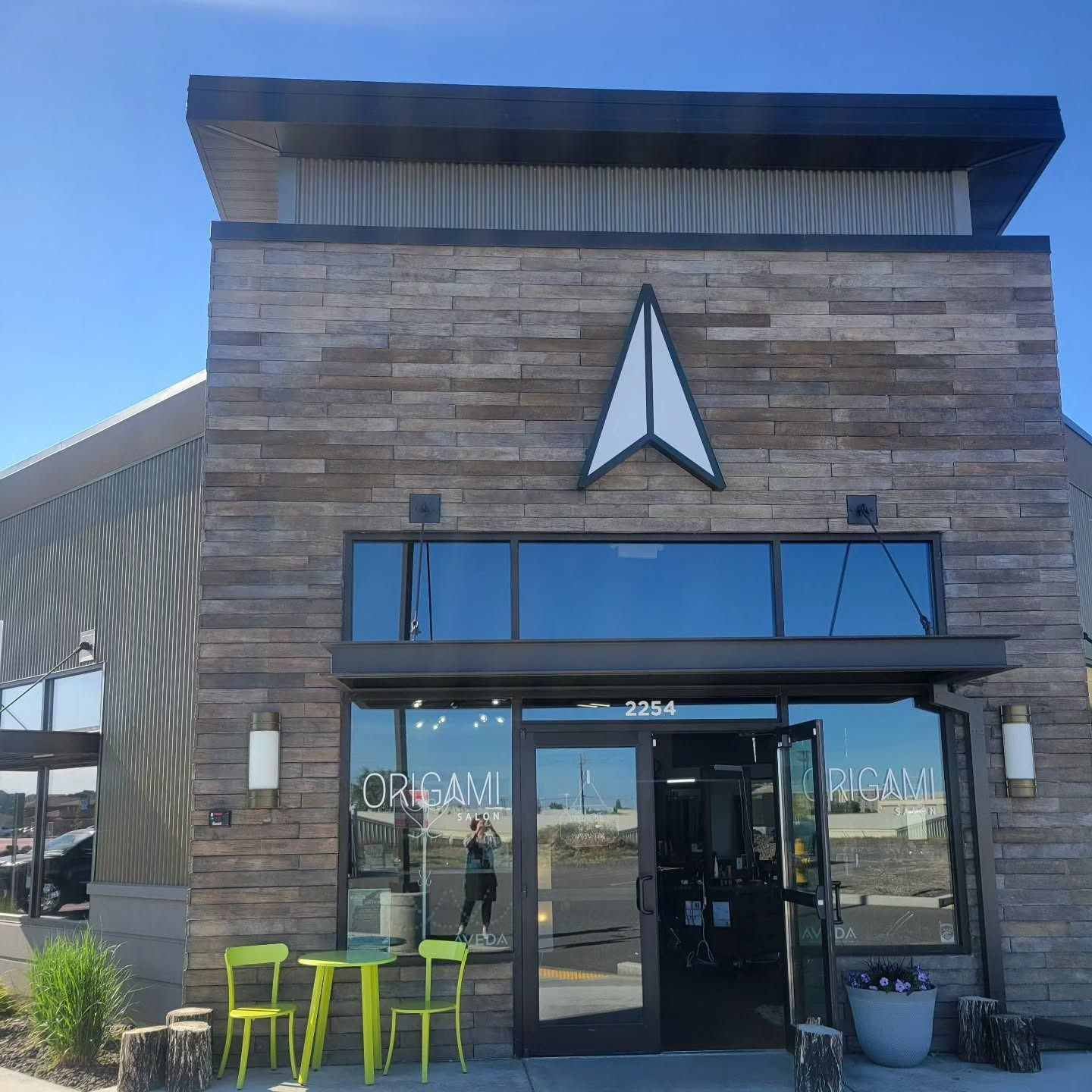 A stone-faced building entrance with a geometric arrow sign above the glass door and a small bistro set on the patio.