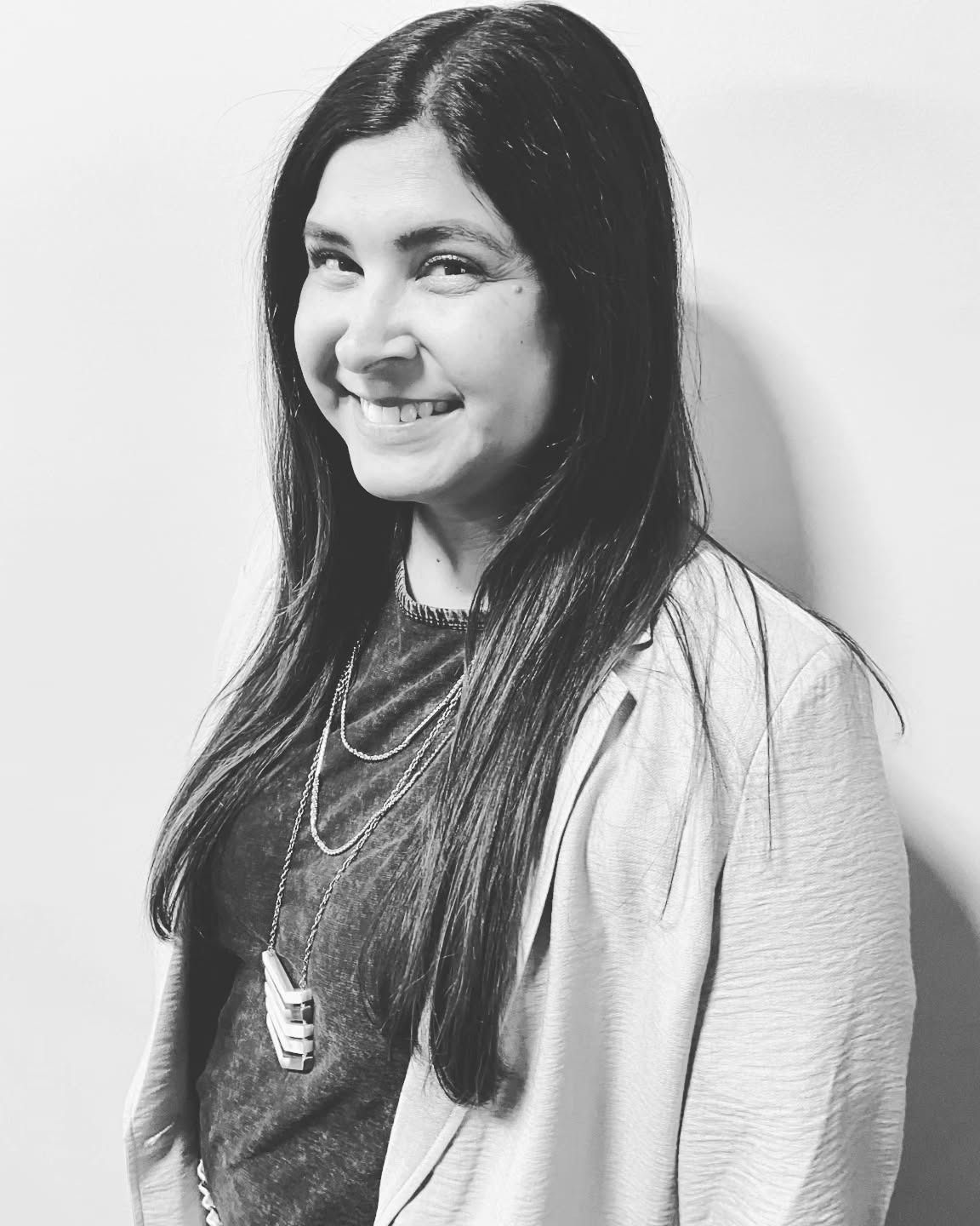 A person with long dark hair smiles while wearing a necklace and a blazer against a plain light wall in black and white.