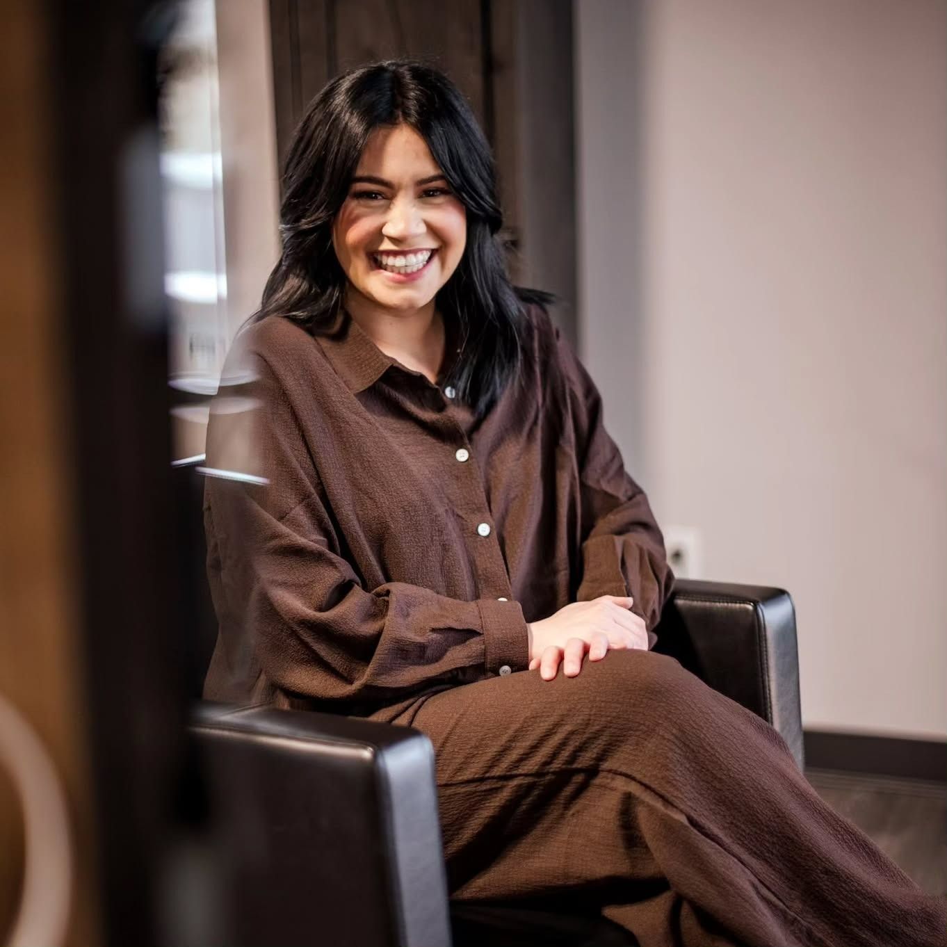 A smiling person with dark hair wears a brown long-sleeved shirt and pants while sitting in a chair in a professional space.