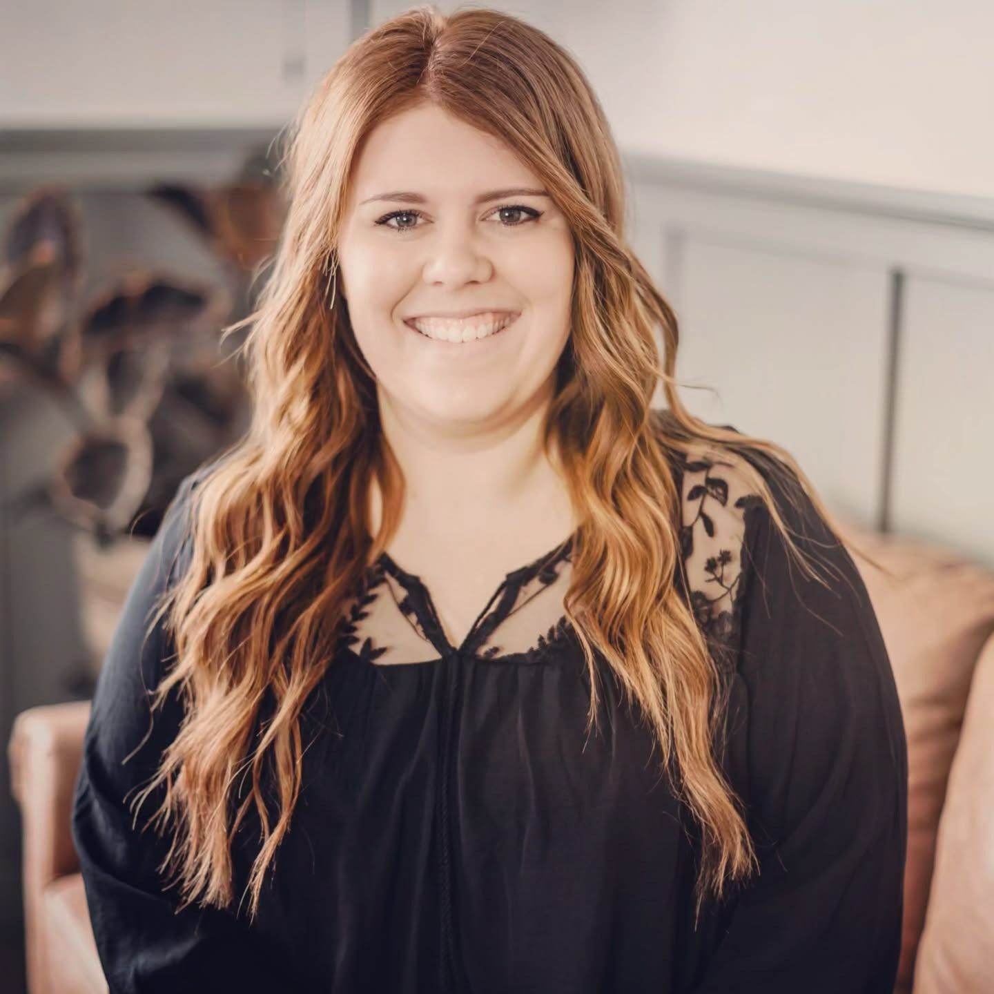Smiling person with long, wavy auburn hair wearing a black long-sleeved shirt with lace detail, sitting indoors.