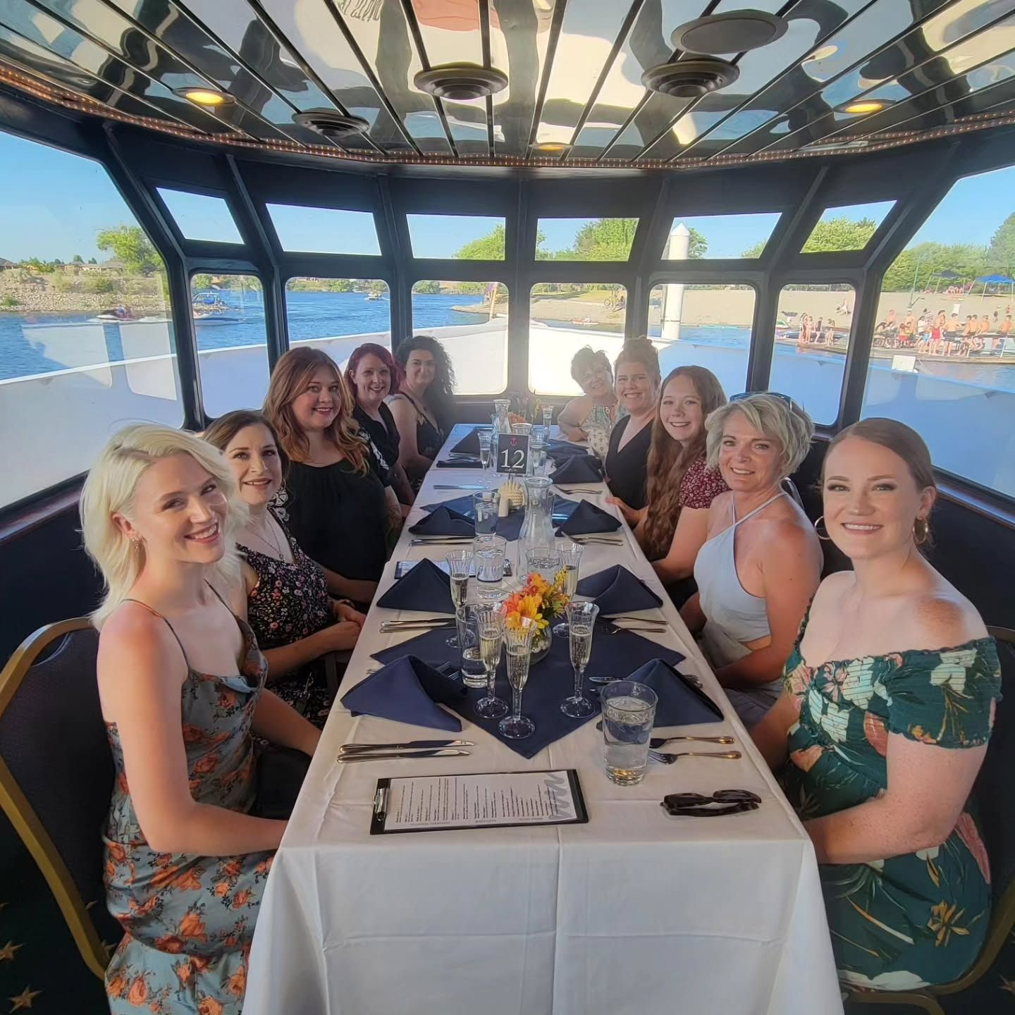 A group of women dining at a table on a boat, with large windows overlooking a river and shoreline.