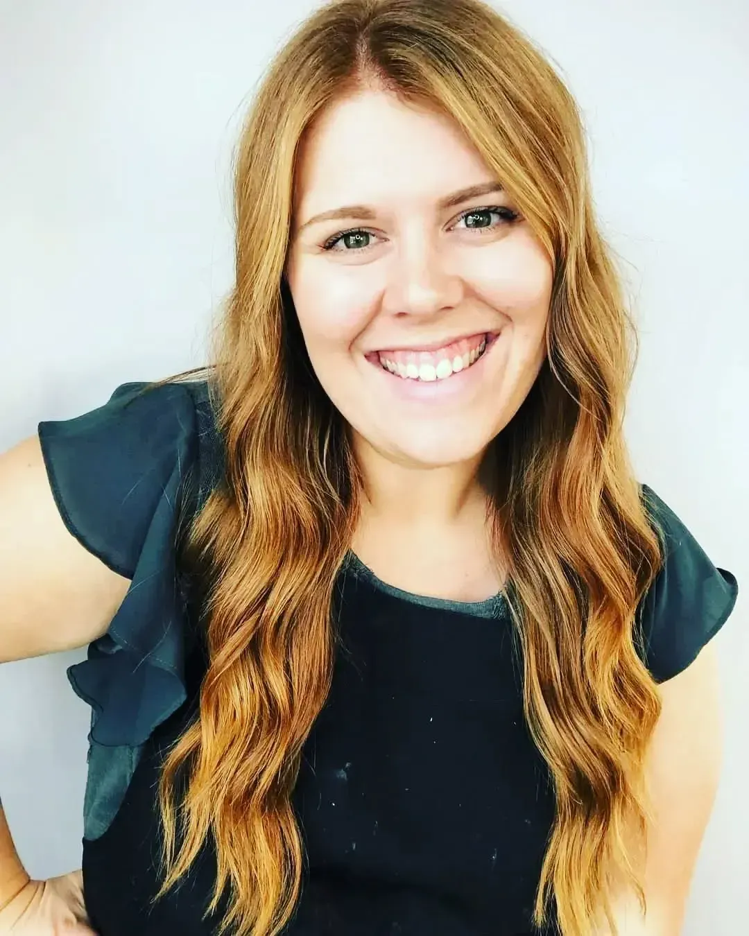 A person with long, wavy blonde hair smiles at the camera, wearing a dark green flutter-sleeve top against a grey wall.