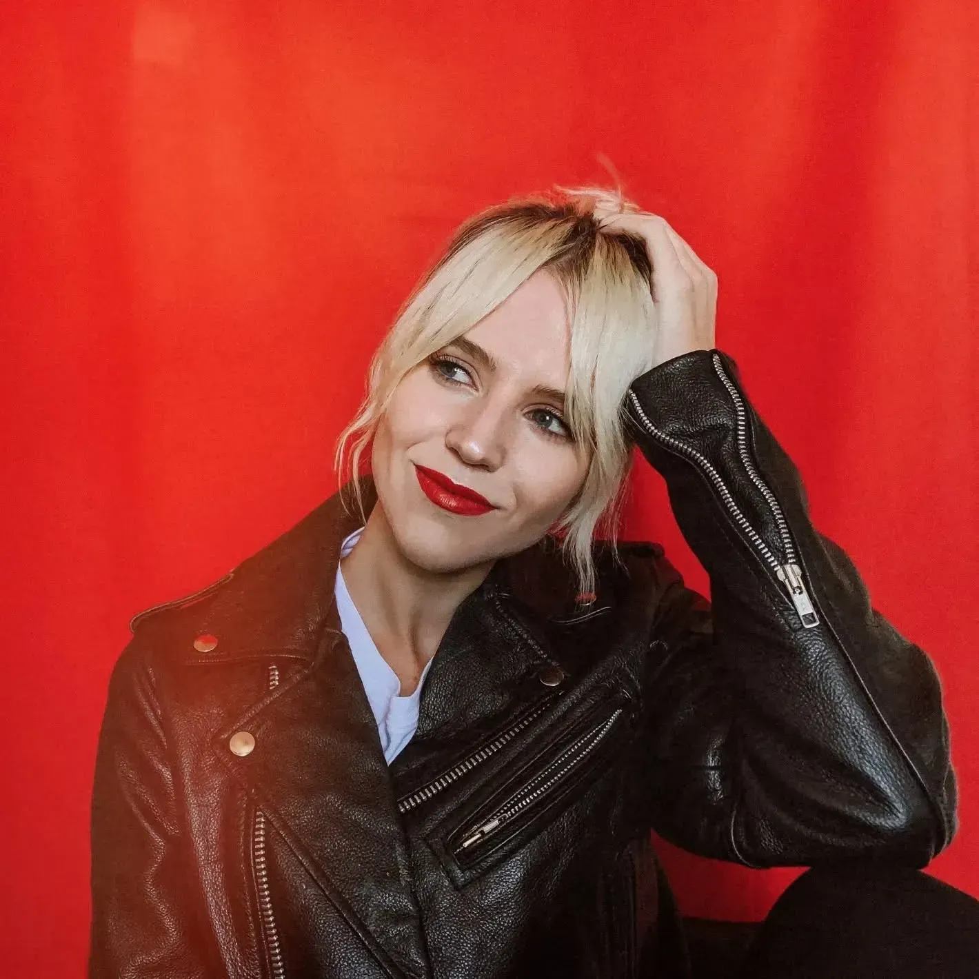 A person with blonde hair and red lipstick poses in a black leather jacket against a bright red background.