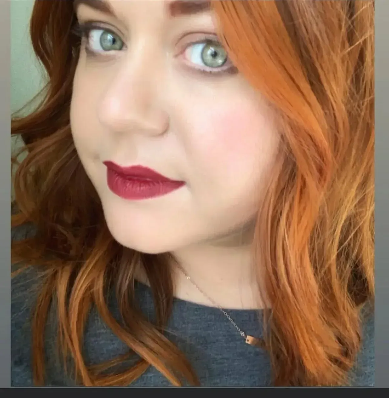 A close-up selfie of a person with wavy, reddish-orange hair, green eyes, and dark berry-colored lipstick.