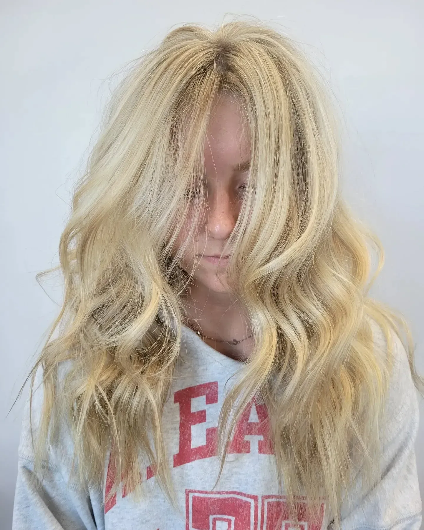 A person with long, blonde, wavy hair that partially obscures their face, wearing a grey sweatshirt with red lettering.