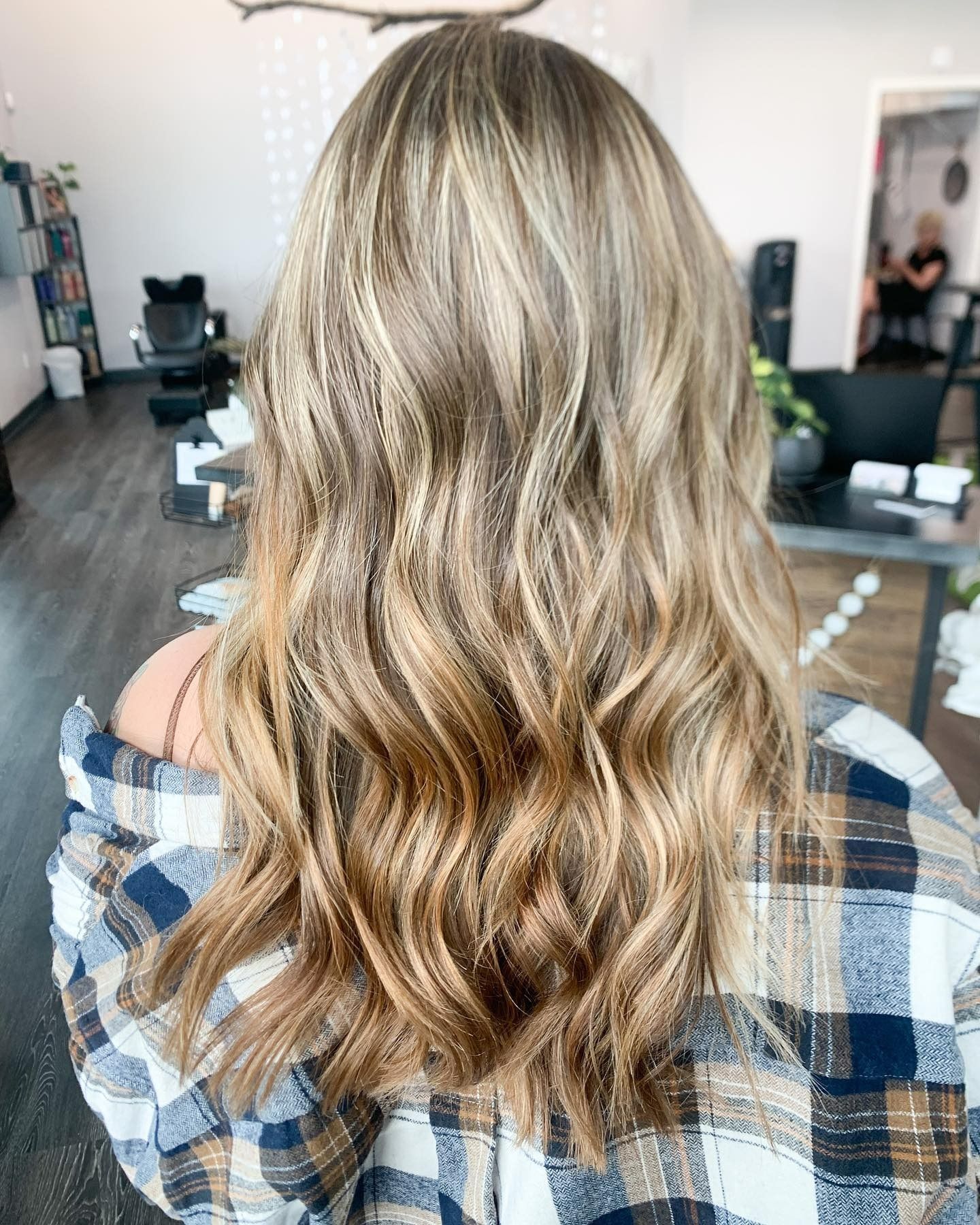 Back view of long, wavy hair with blonde balayage highlights, worn by a person in a plaid flannel shirt in a salon.