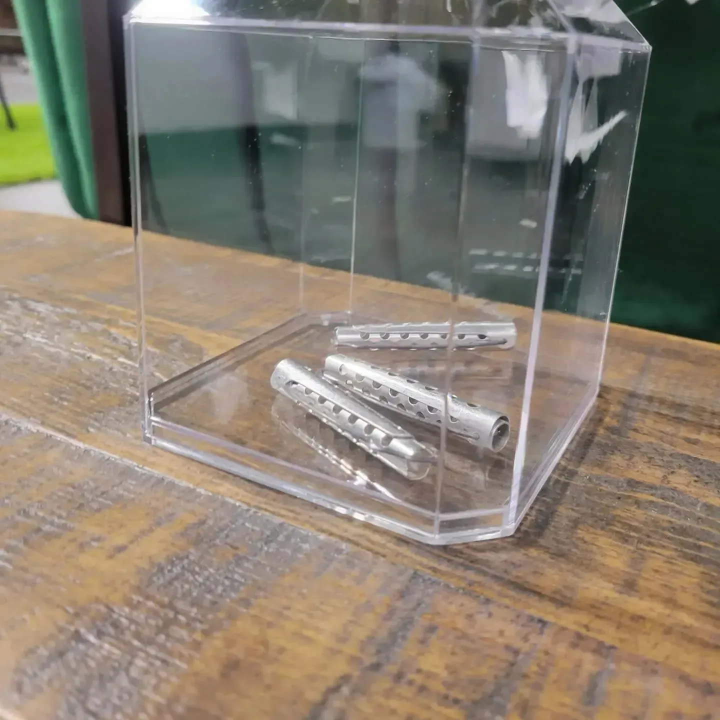 Three metal wall anchors inside a clear plastic display cube, placed on a wooden table.