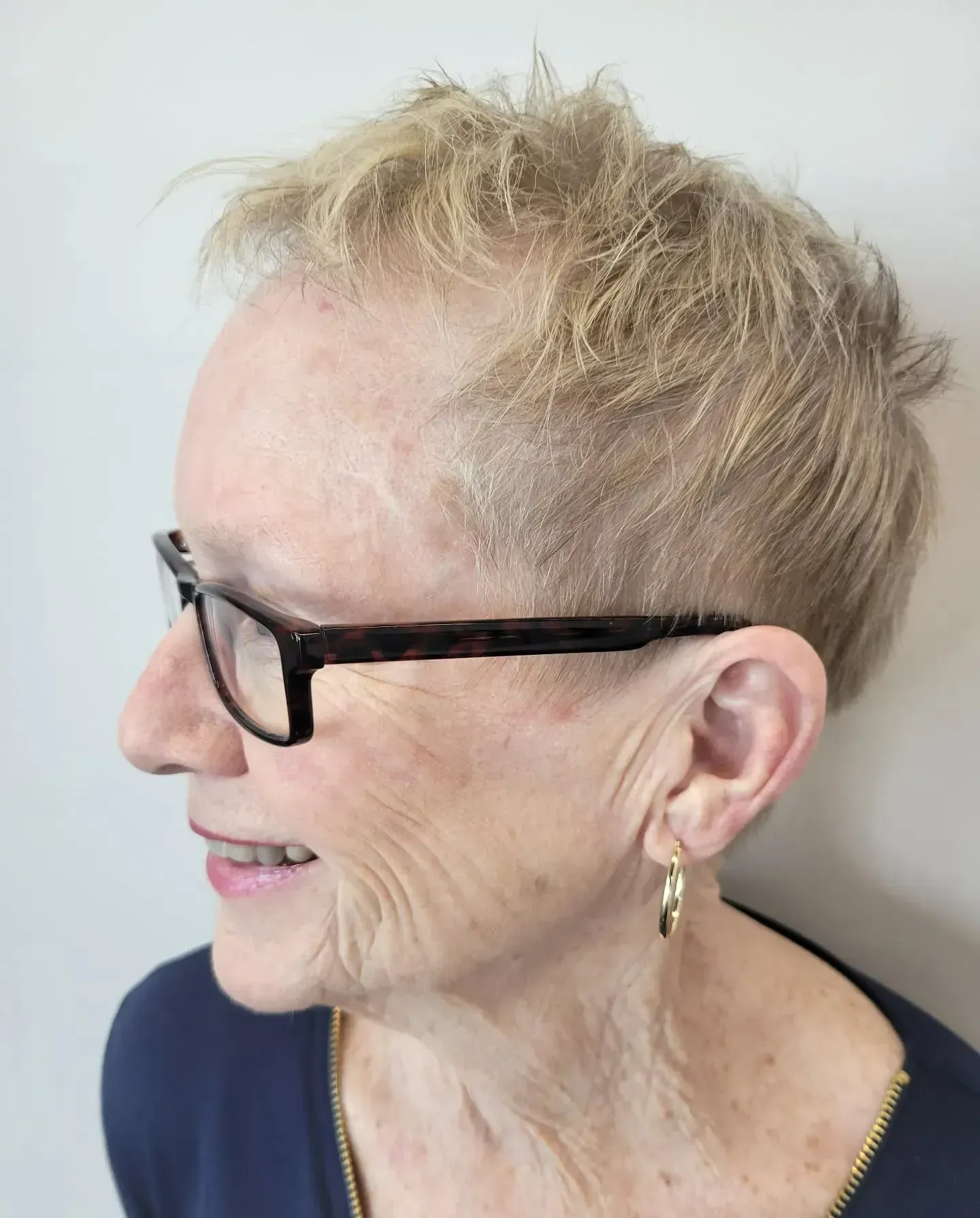 A profile view of a person with short, light-colored hair wearing dark-framed glasses and small gold hoop earrings.