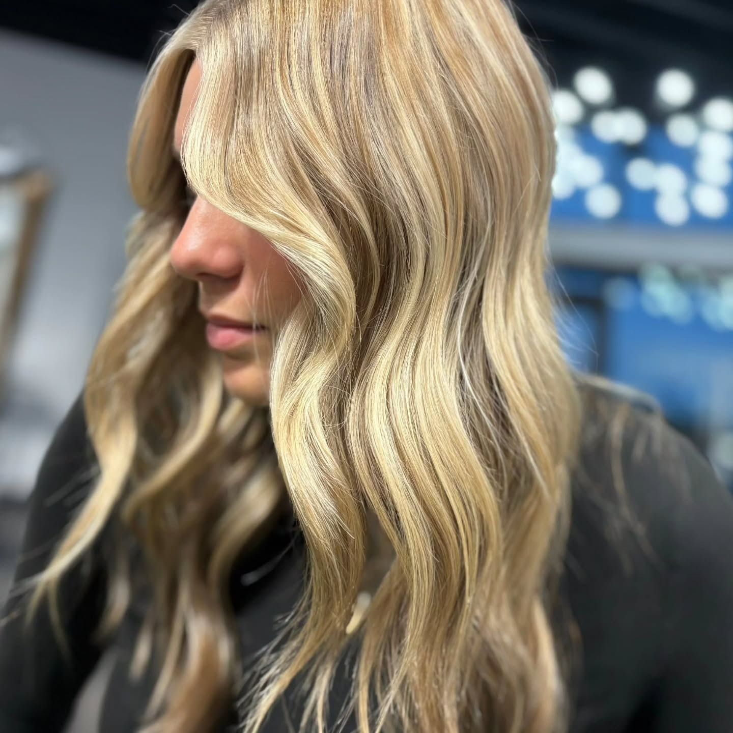 A person with long, wavy blonde hair styled with bright highlights, viewed from the side in a salon setting.