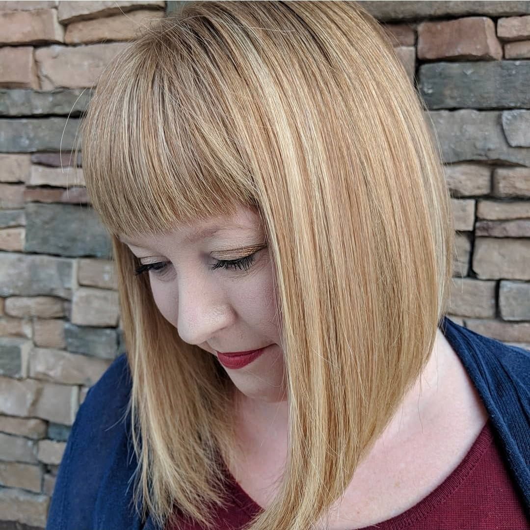 A woman with crimped, shoulder-length blonde hair and bangs looks down against a stone wall background.