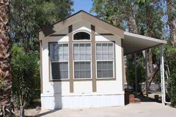 Small white and brown mobile home with covered carport, set among trees.