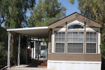 Tan and white cabin with a carport, set against a backdrop of trees.