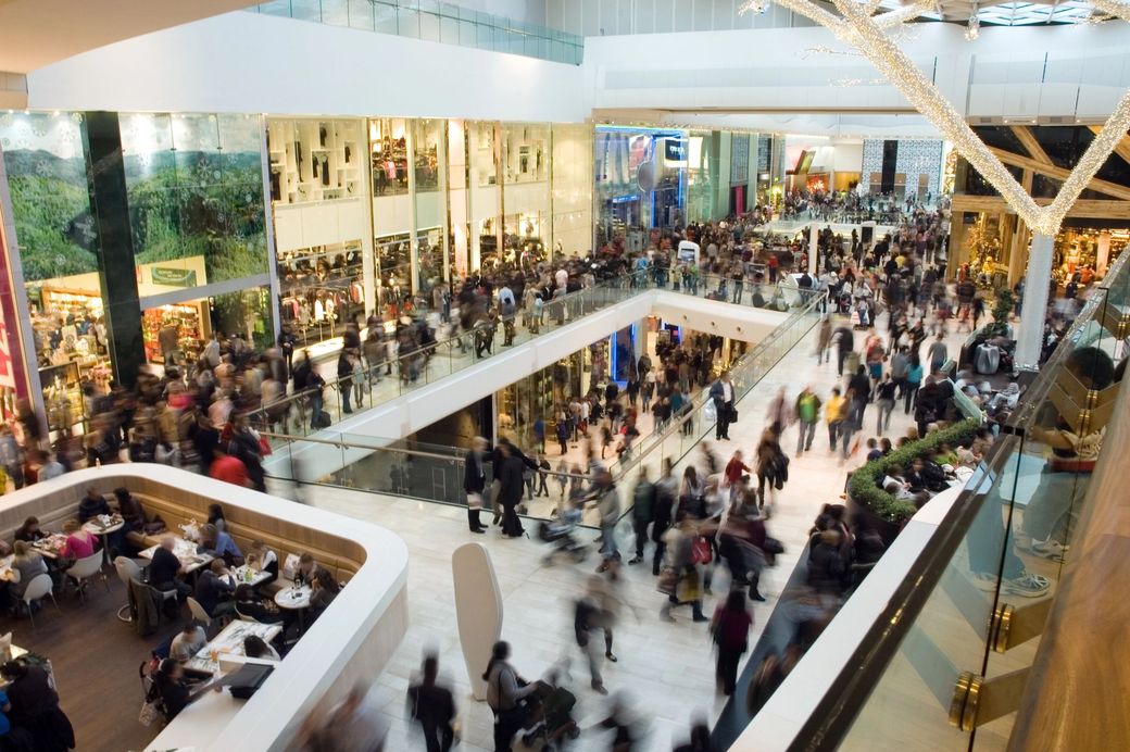 Interno di un centro commerciale affollato, con numerose persone sfocate che si muovono tra negozi e scale mobili.