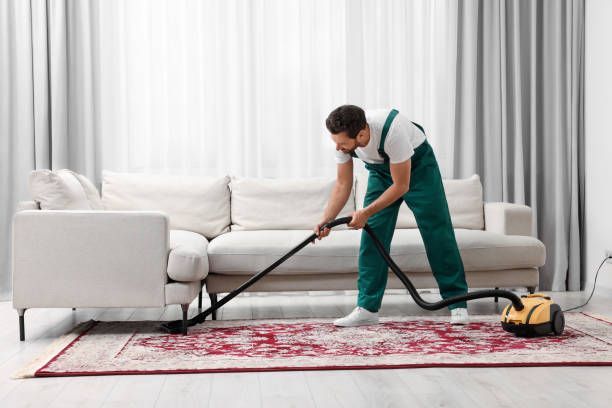 Man wearing an apron and red bandana vacuuming a rug in a living room.
