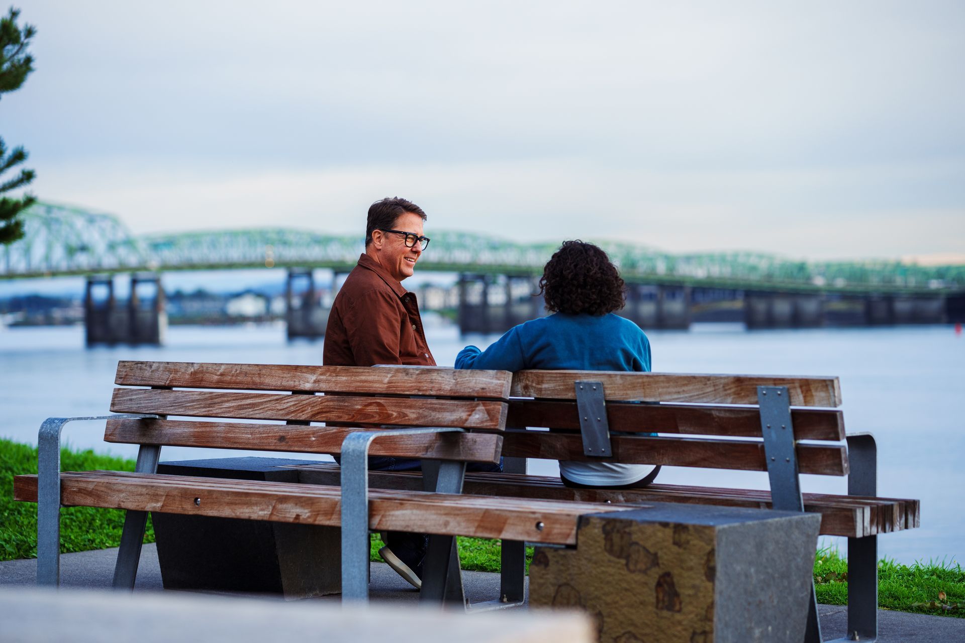 Two people sit on a wooden bench overlooking a wide river with a long metal bridge in the background under a cloudy sky.