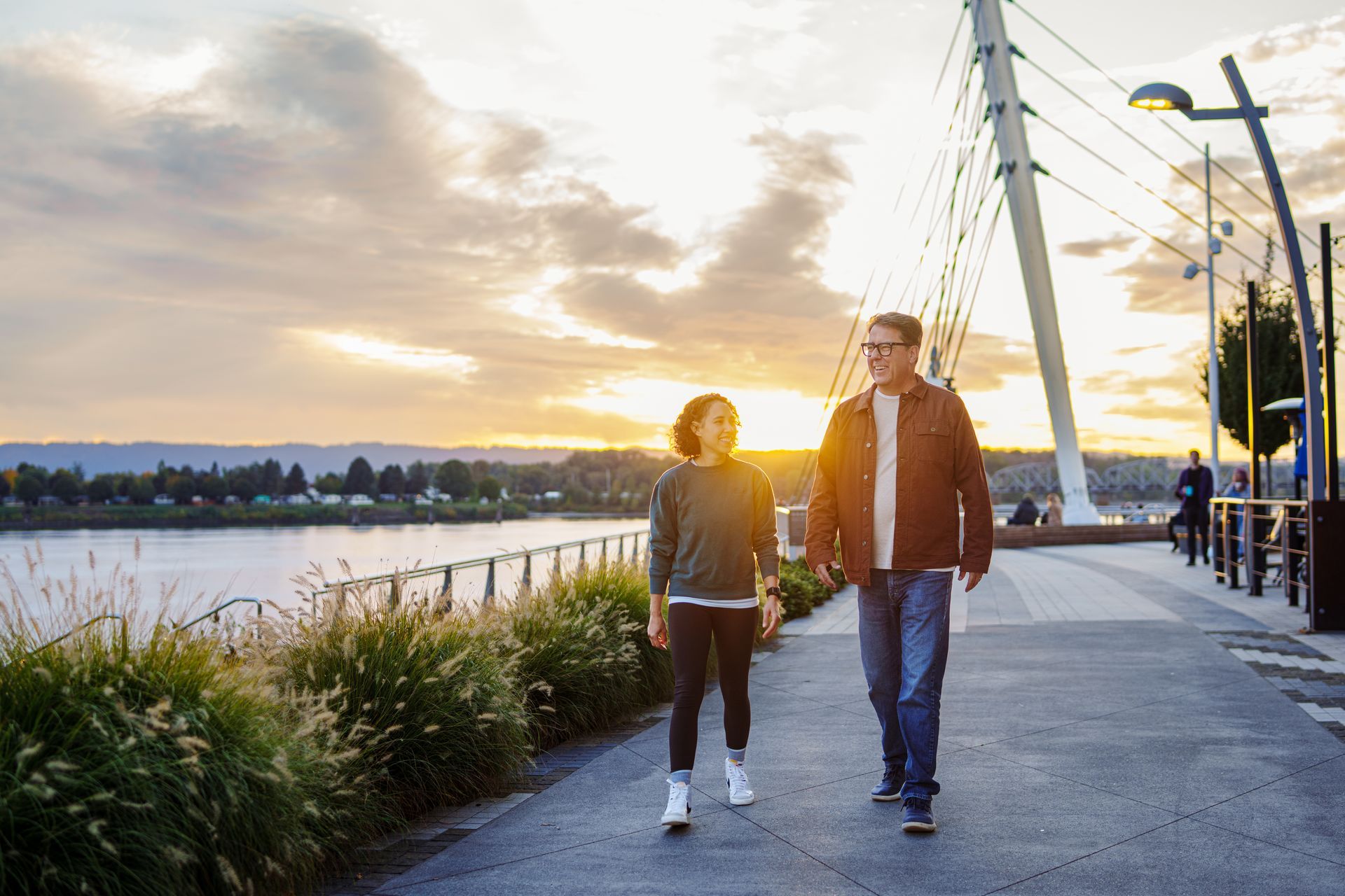 Two people walk together along a paved waterfront path during sunset, with a bridge structure visible in the background.