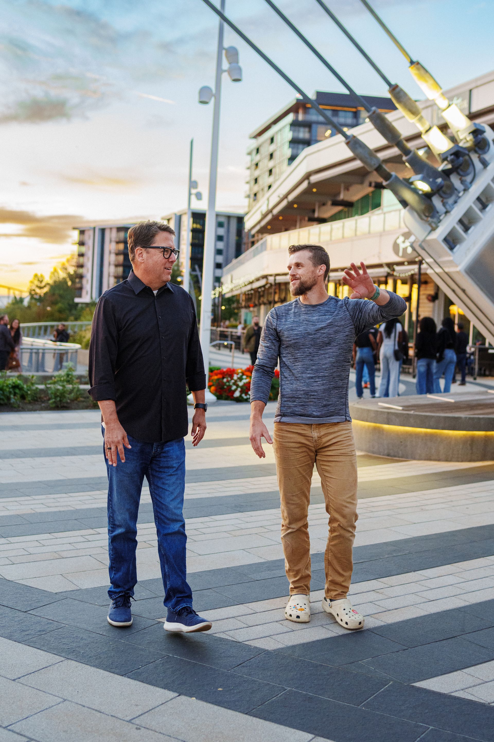 Two men walk outdoors on a paved plaza, one gesturing as they converse, with modern buildings and structural cables nearby.