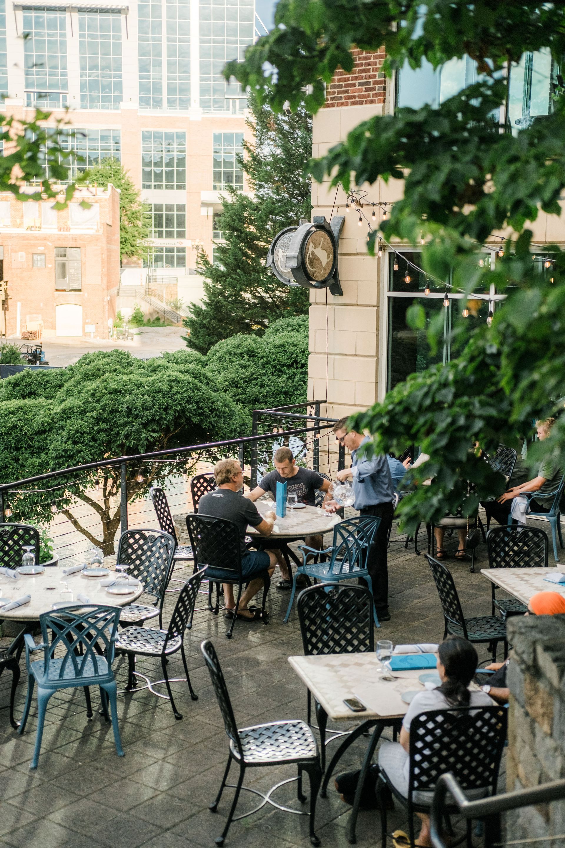 Outdoor café patio with black metal tables and chairs, diners under a leafy tree and city buildings behind