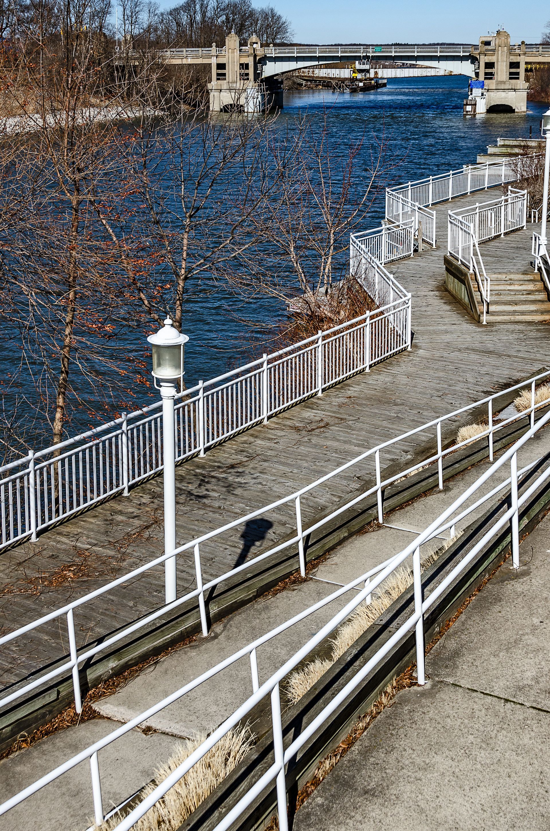 Riverside walkway with white railings beside blue water and a bridge in the distance