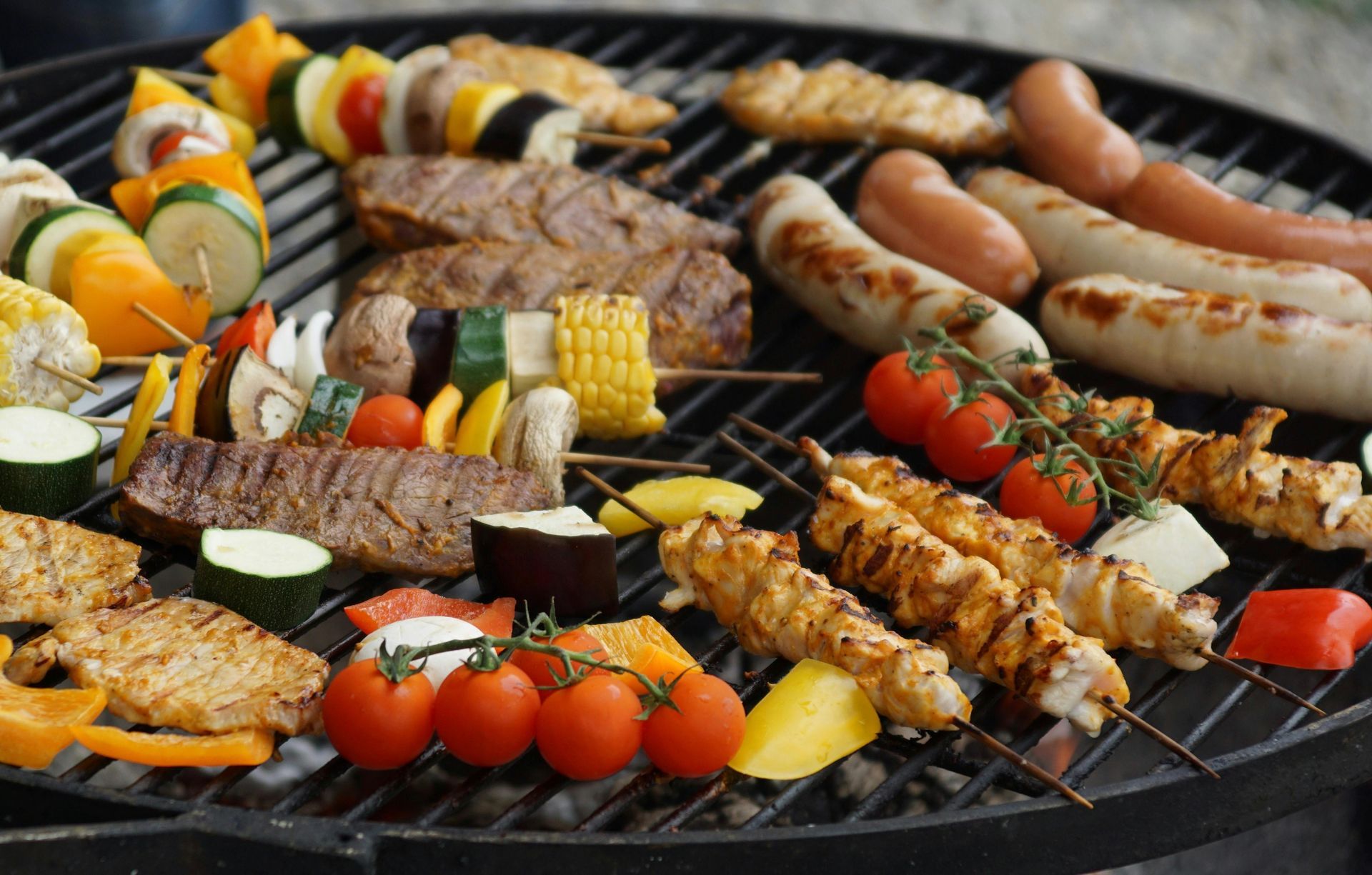 Assorted meat, sausage, and vegetable skewers grilling over hot coals on a barbecue.