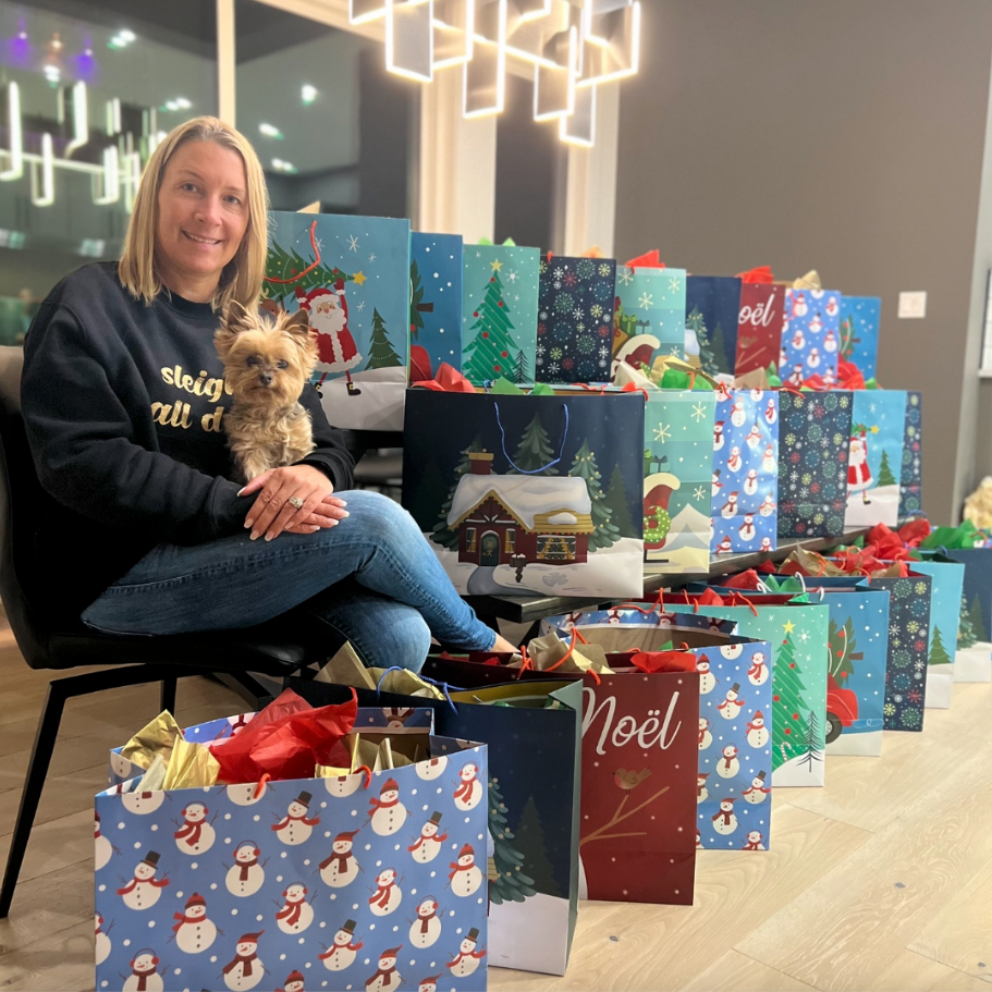 A woman is sitting in a chair with a dog in front of a stack of christmas bags that say noel