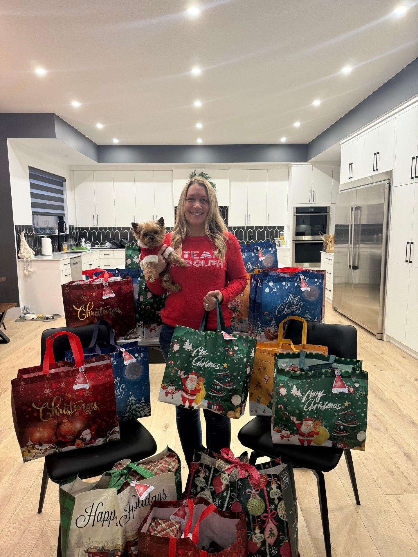 A woman is standing in a kitchen holding a dog and a bunch of christmas bags.