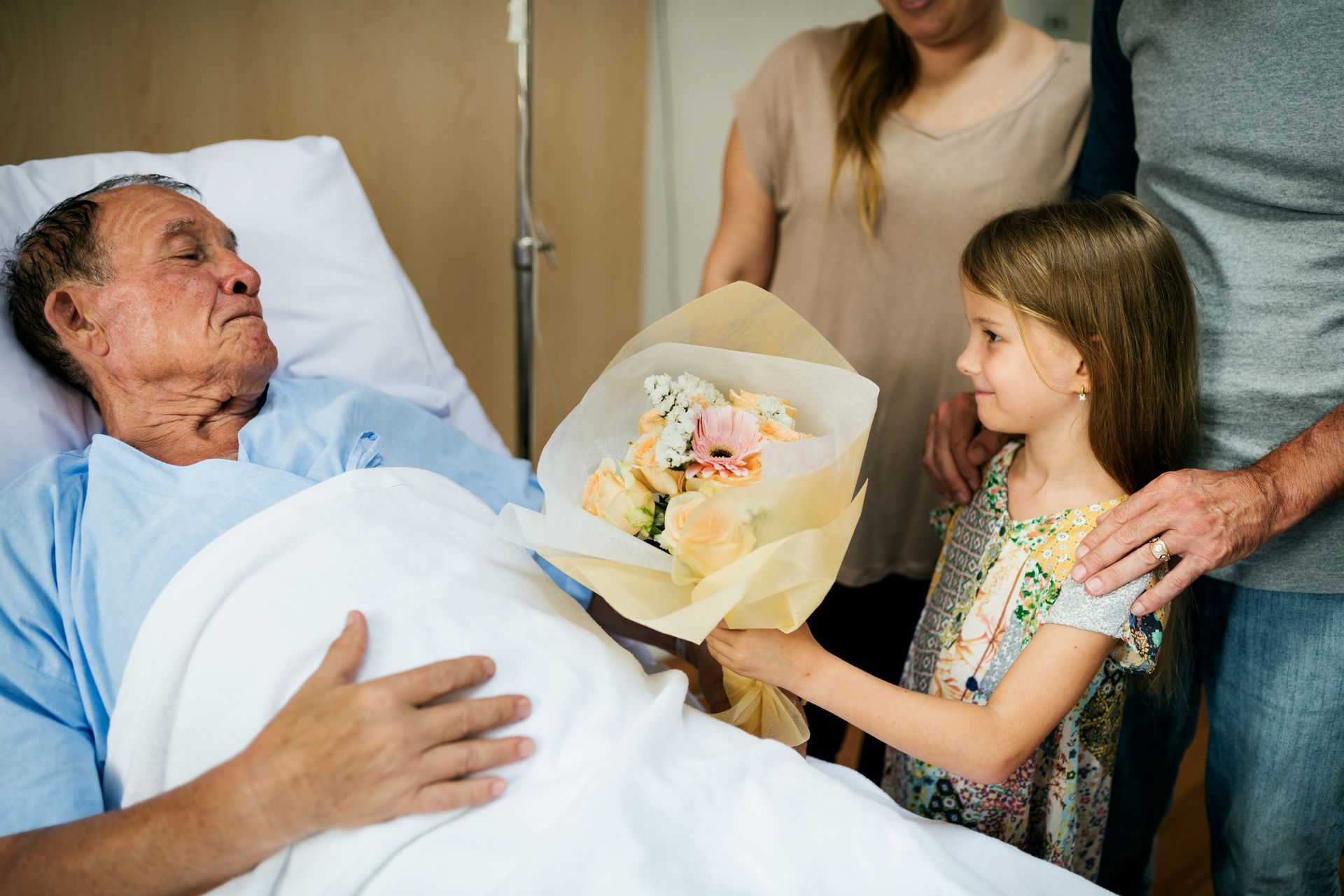 Hospital room scene with an elderly patient in bed receiving flowers from a child and family members