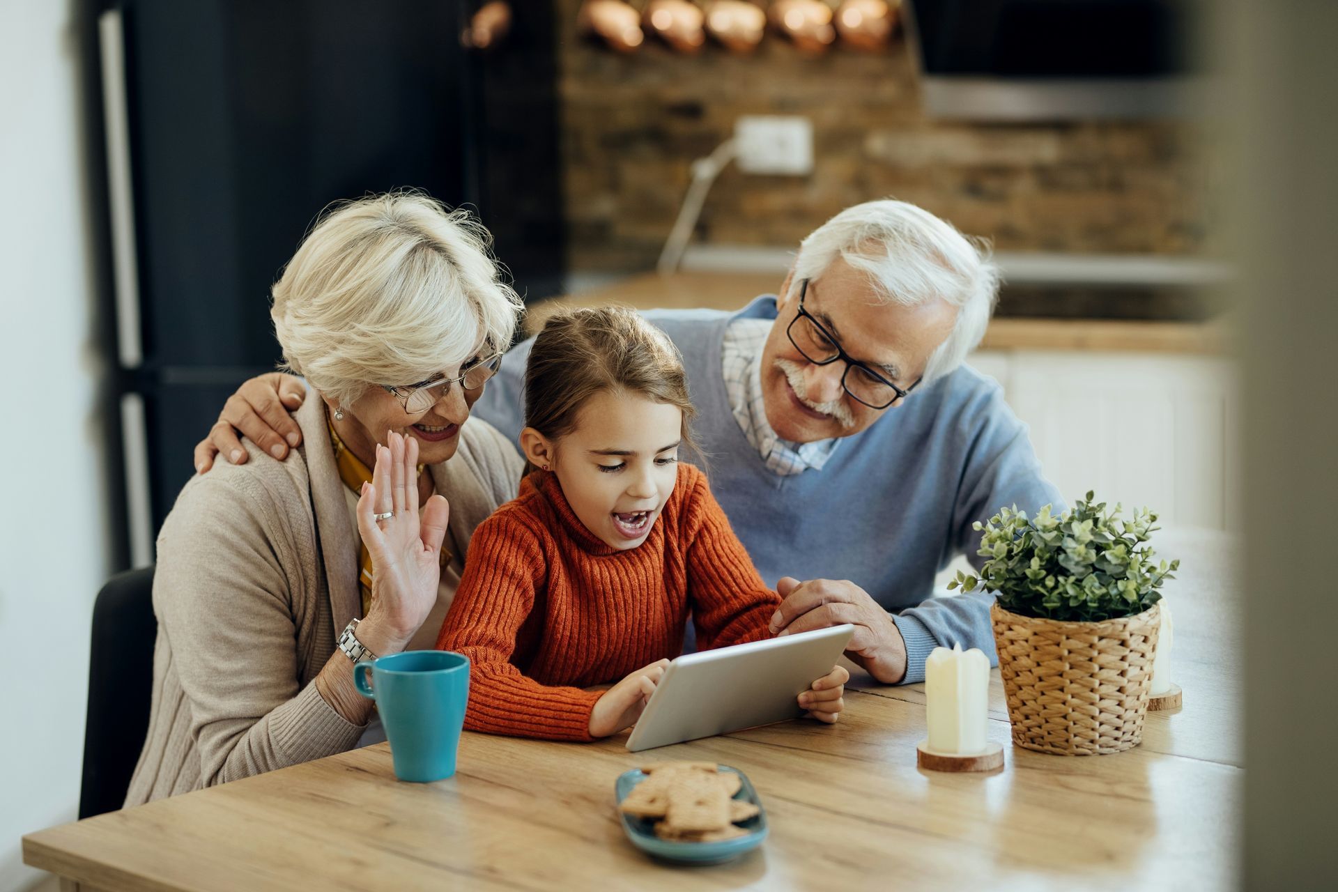 Three people gathered around a tablet at a kitchen table, smiling and leaning in close.