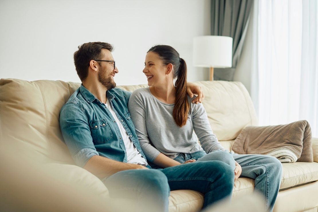 Two people sitting on a beige couch, smiling at each other in a bright living room.