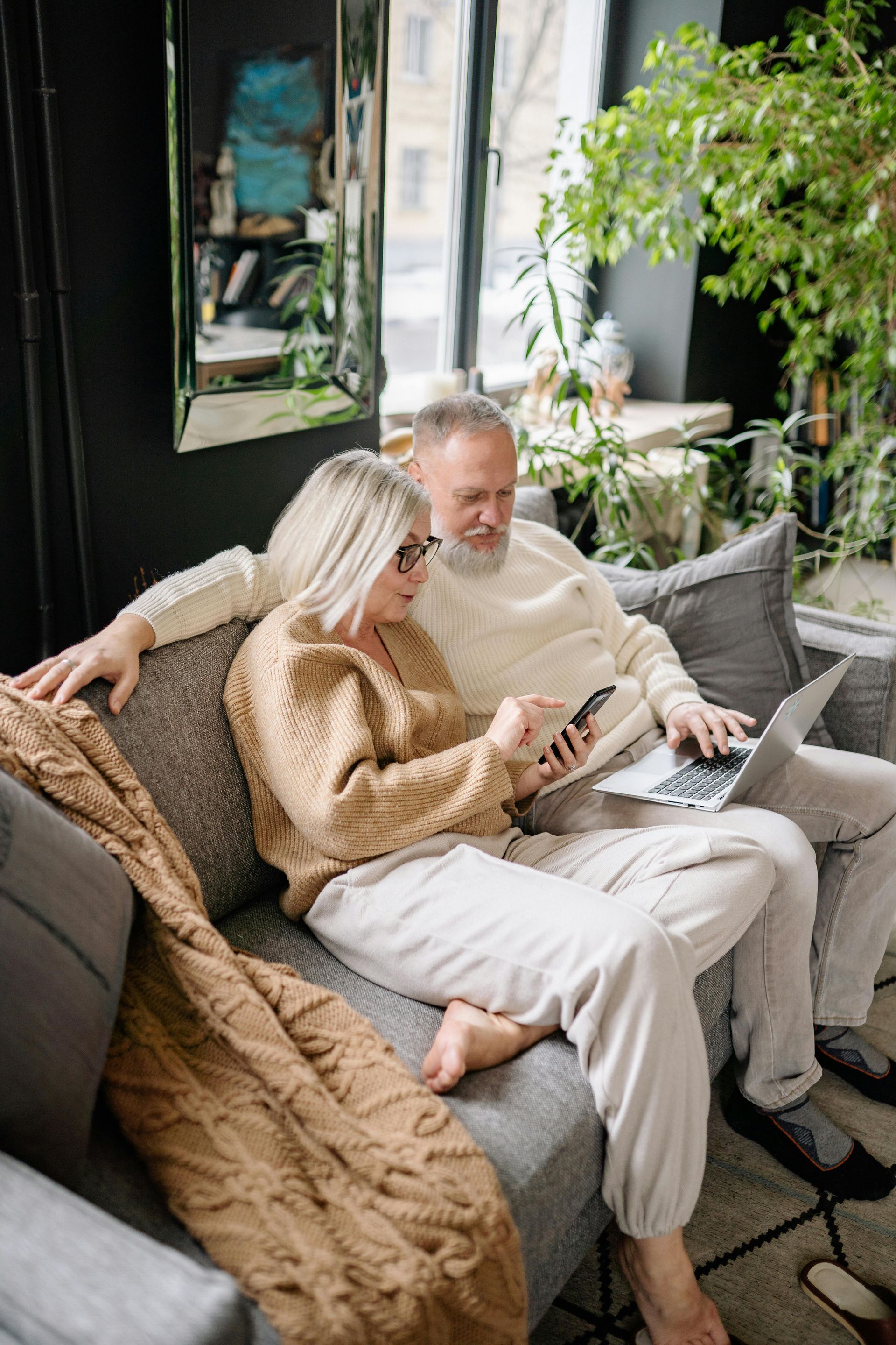 Couple relaxing on a couch, looking at a tablet in a cozy living room with plants.