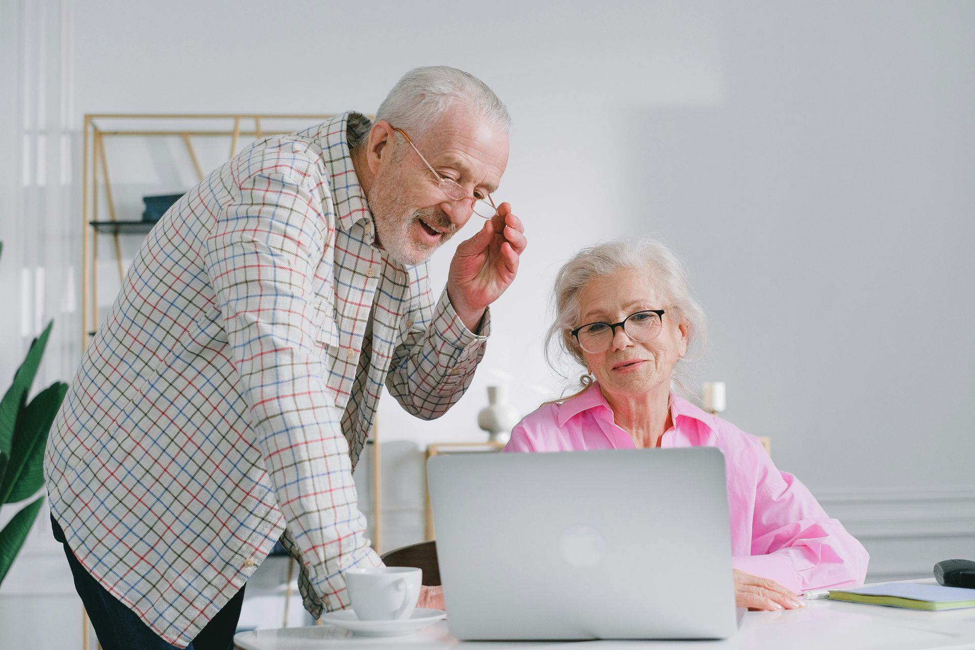 Two older adults working together at a laptop in a bright home office, one leaning in to point at the screen.