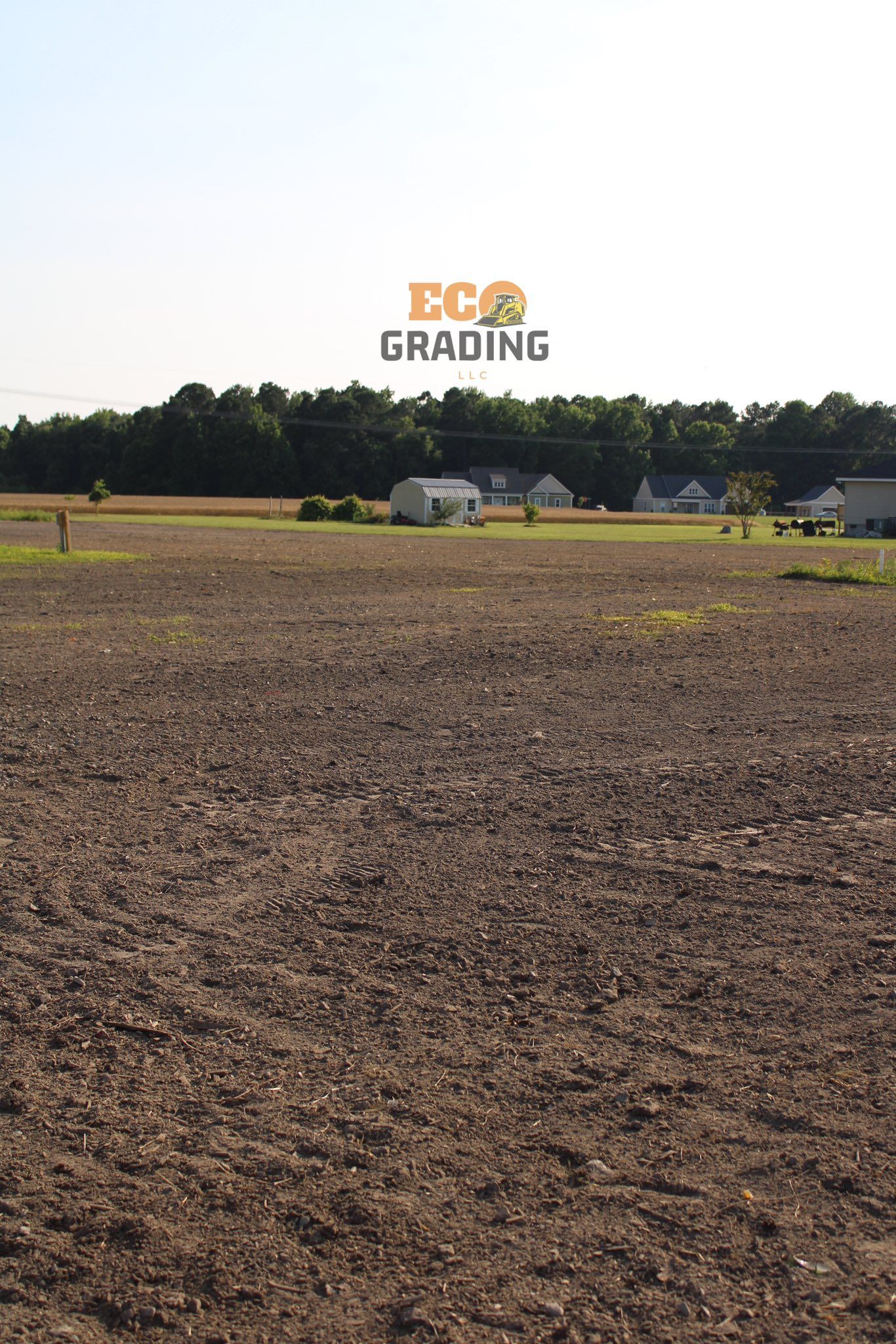 Brown plowed field with trees and houses in the background under a bright sky.