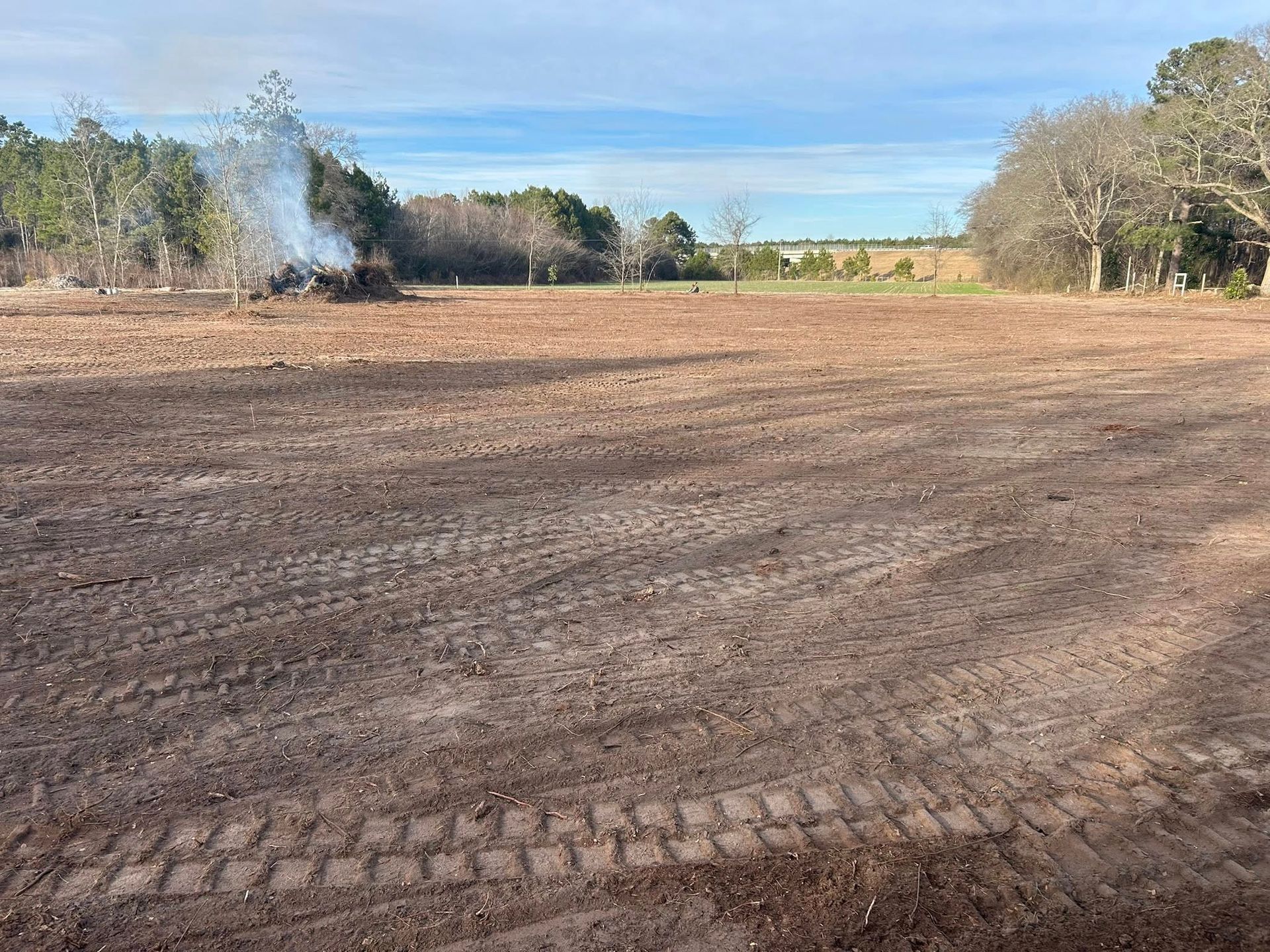 A cleared field with tire tracks. A burning pile of brush is in the distance, under a blue sky.