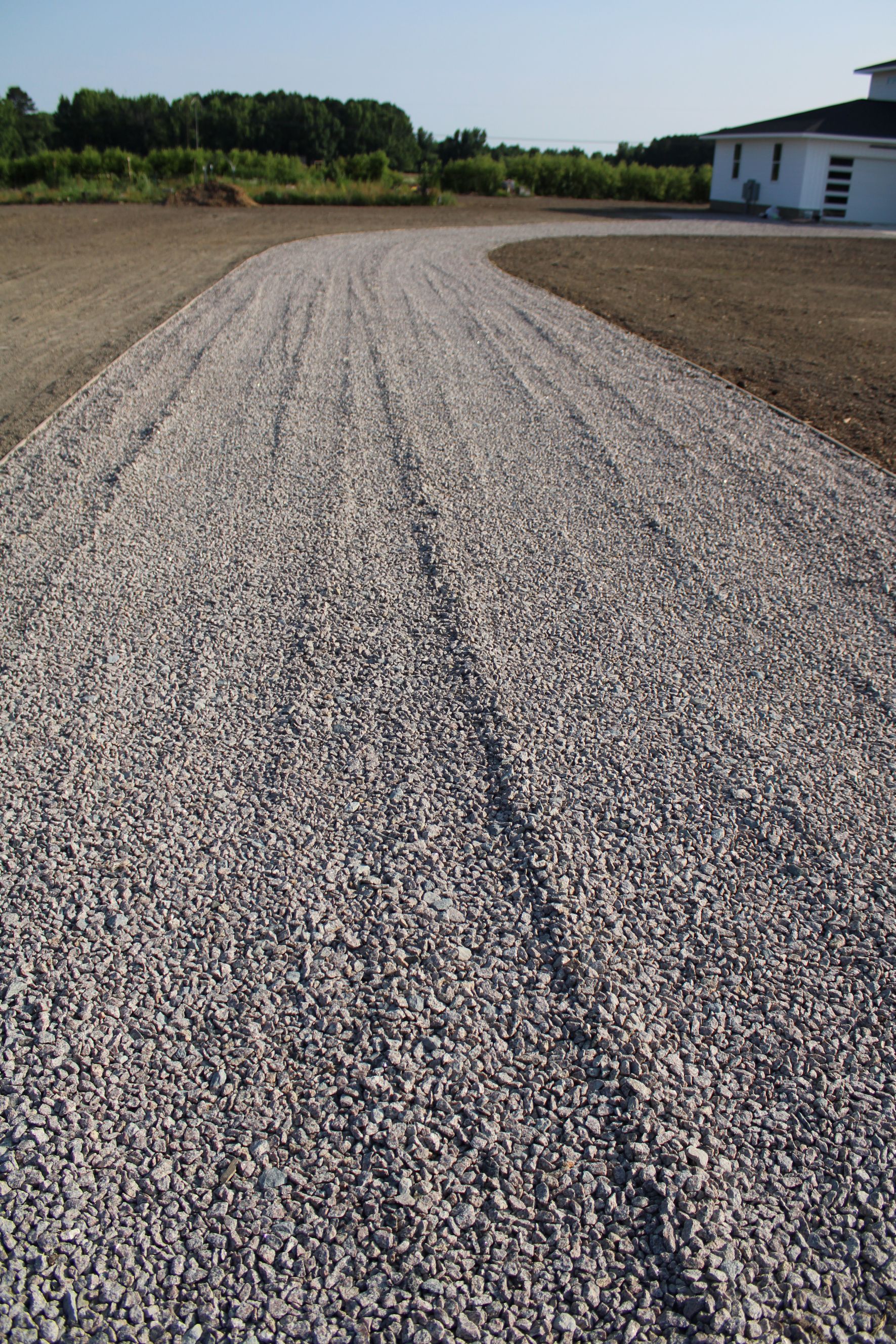 Gravel driveway curving towards a house, brown earth on either side, sunny day.