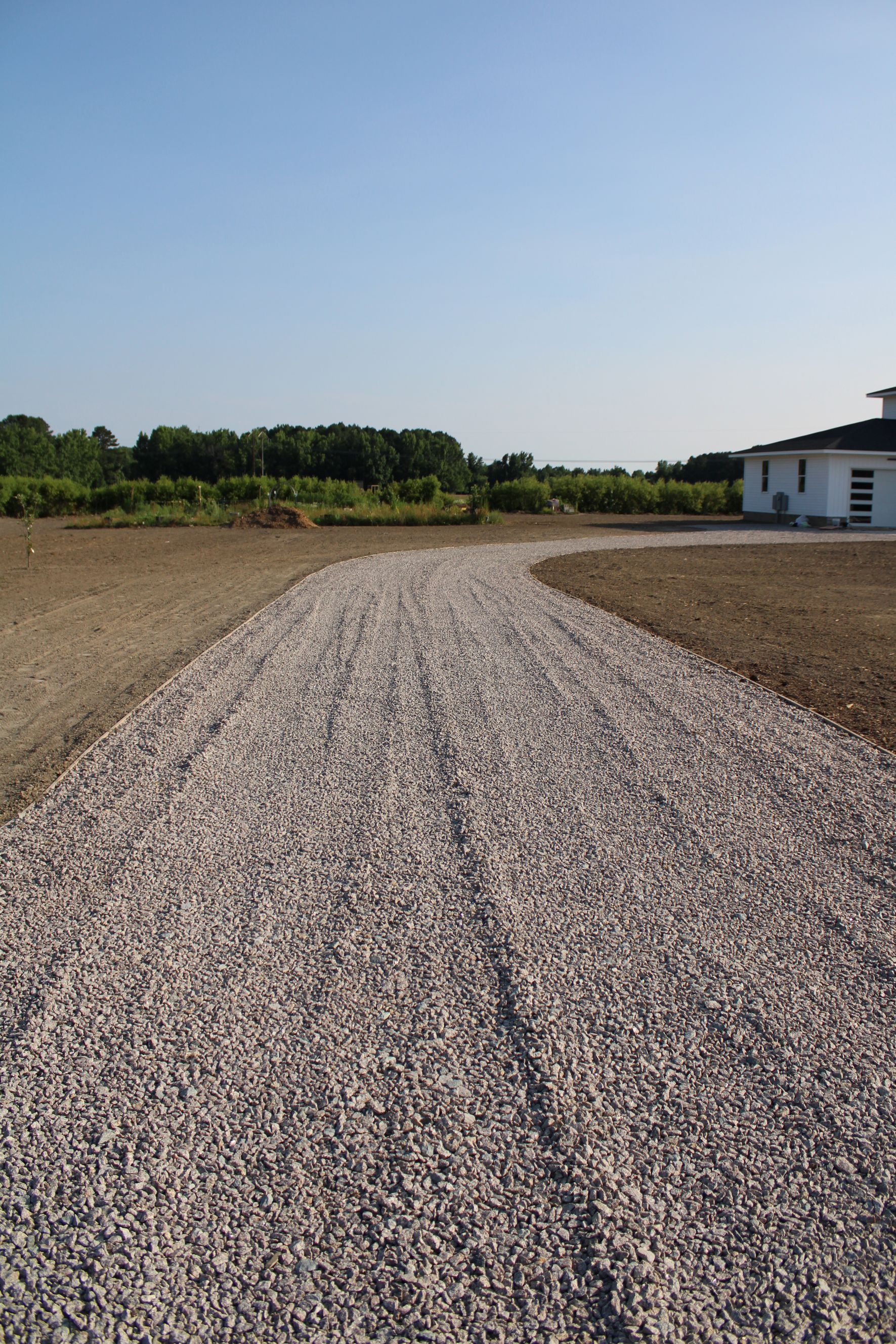 Gravel driveway curves toward white building with trees in the background under a blue sky.