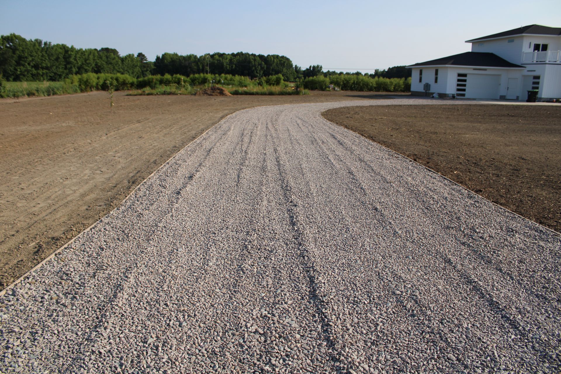 A gravel driveway curves towards a white house, surrounded by bare earth and a tree line.