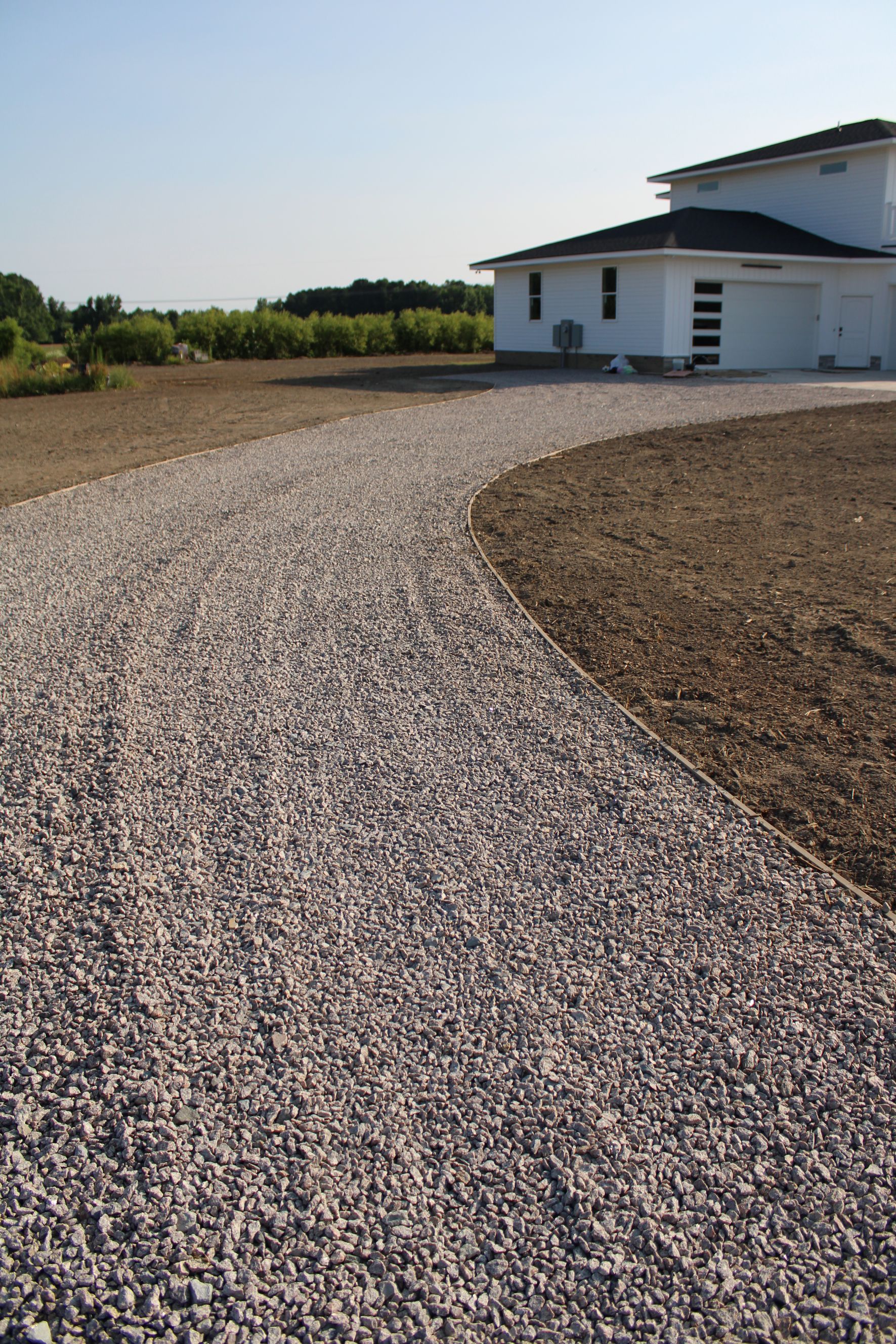 Gravel driveway curves toward a white house with a black roof and a clear blue sky.