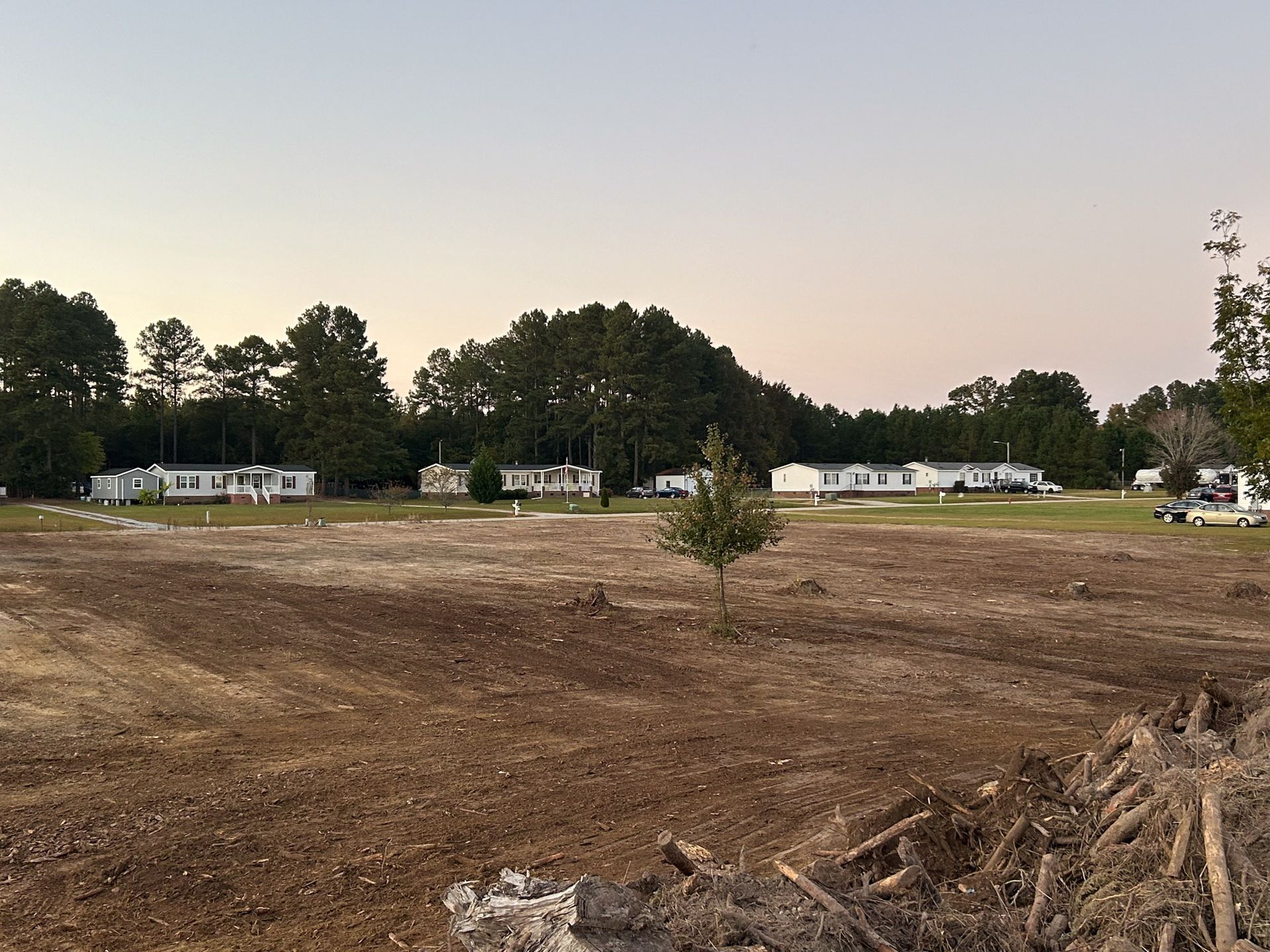 Cleared dirt foreground with mobile homes in background under a dusky sky.