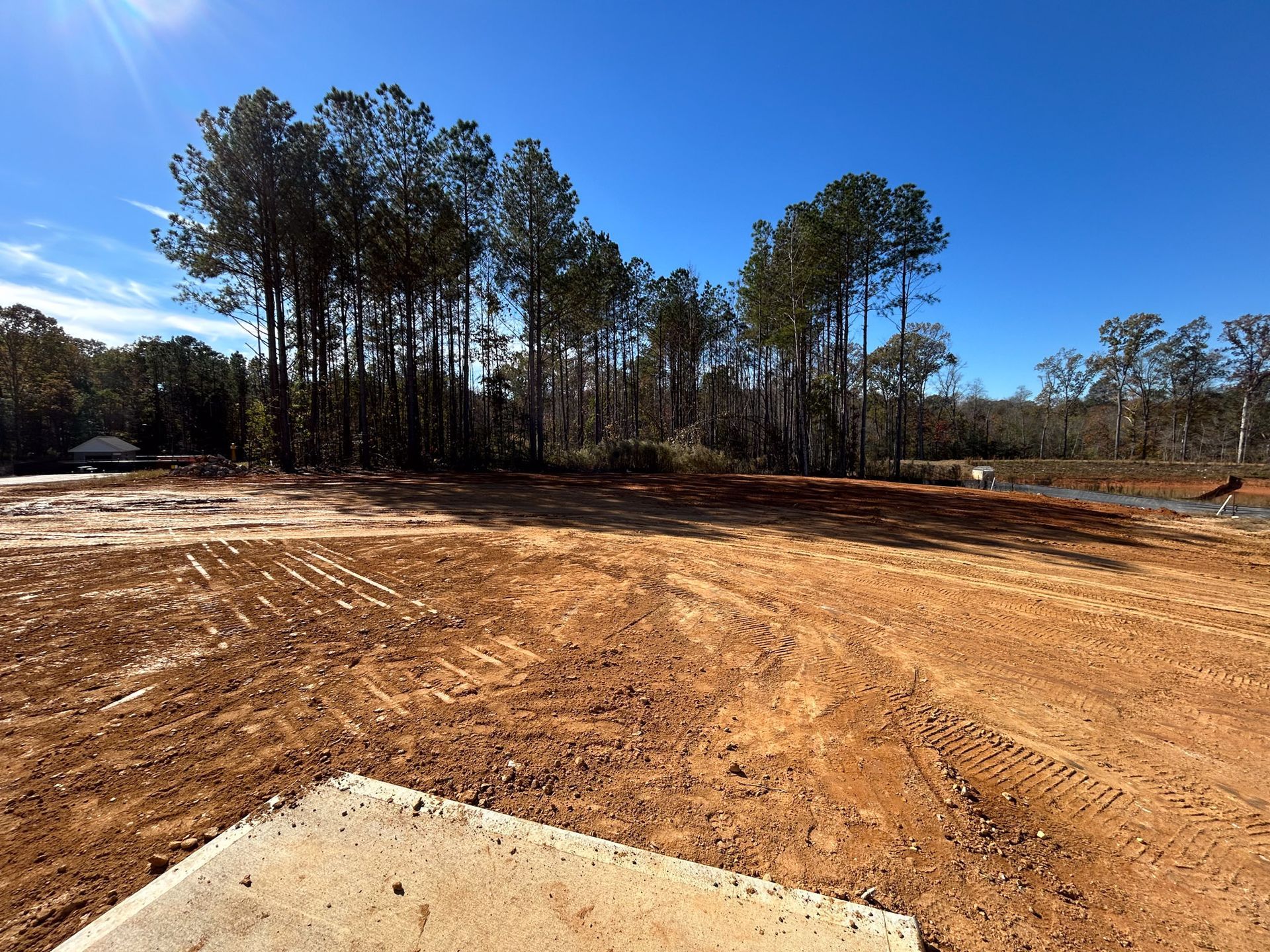 Cleared lot with red dirt, concrete slab, trees, and blue sky.