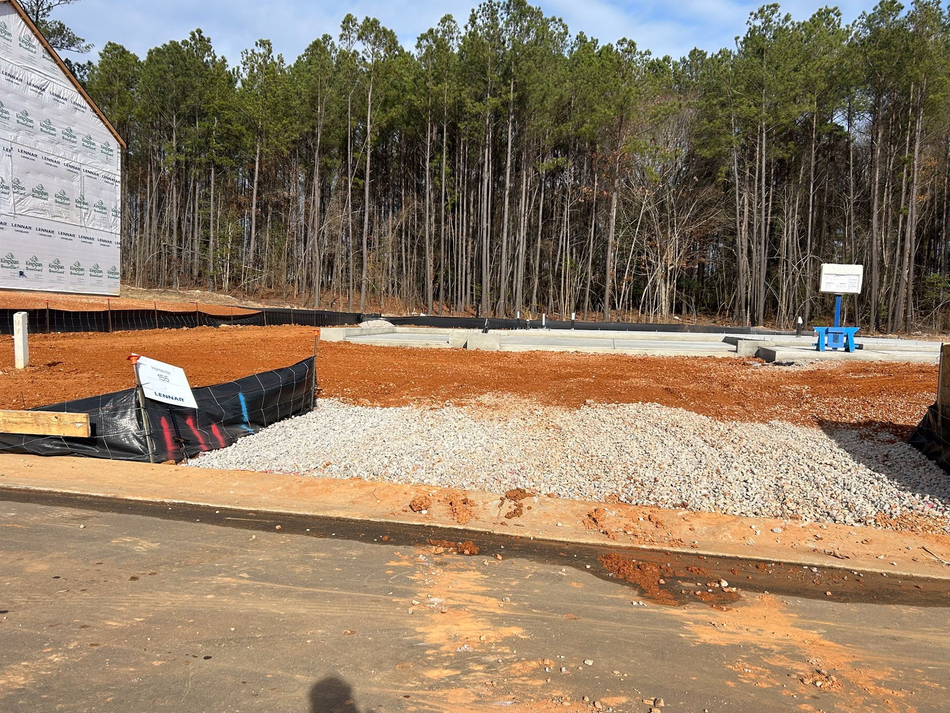 Construction site with gravel, red soil, and trees in the background. A house frame is visible on the left.