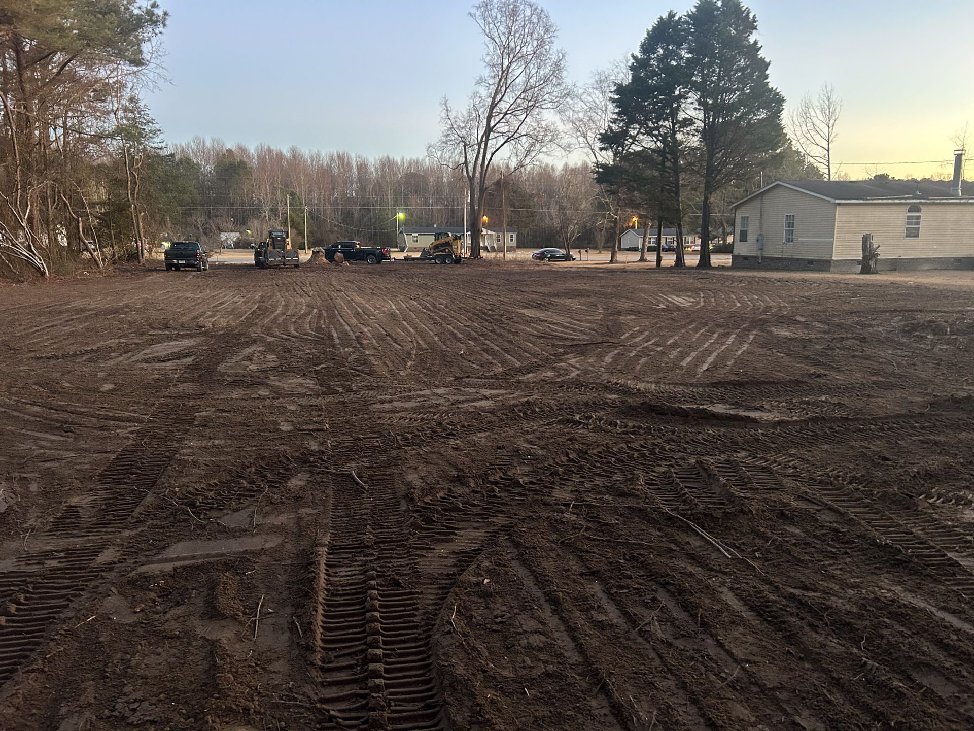 Muddy, leveled construction site with heavy machinery, small buildings, and trees in the background.