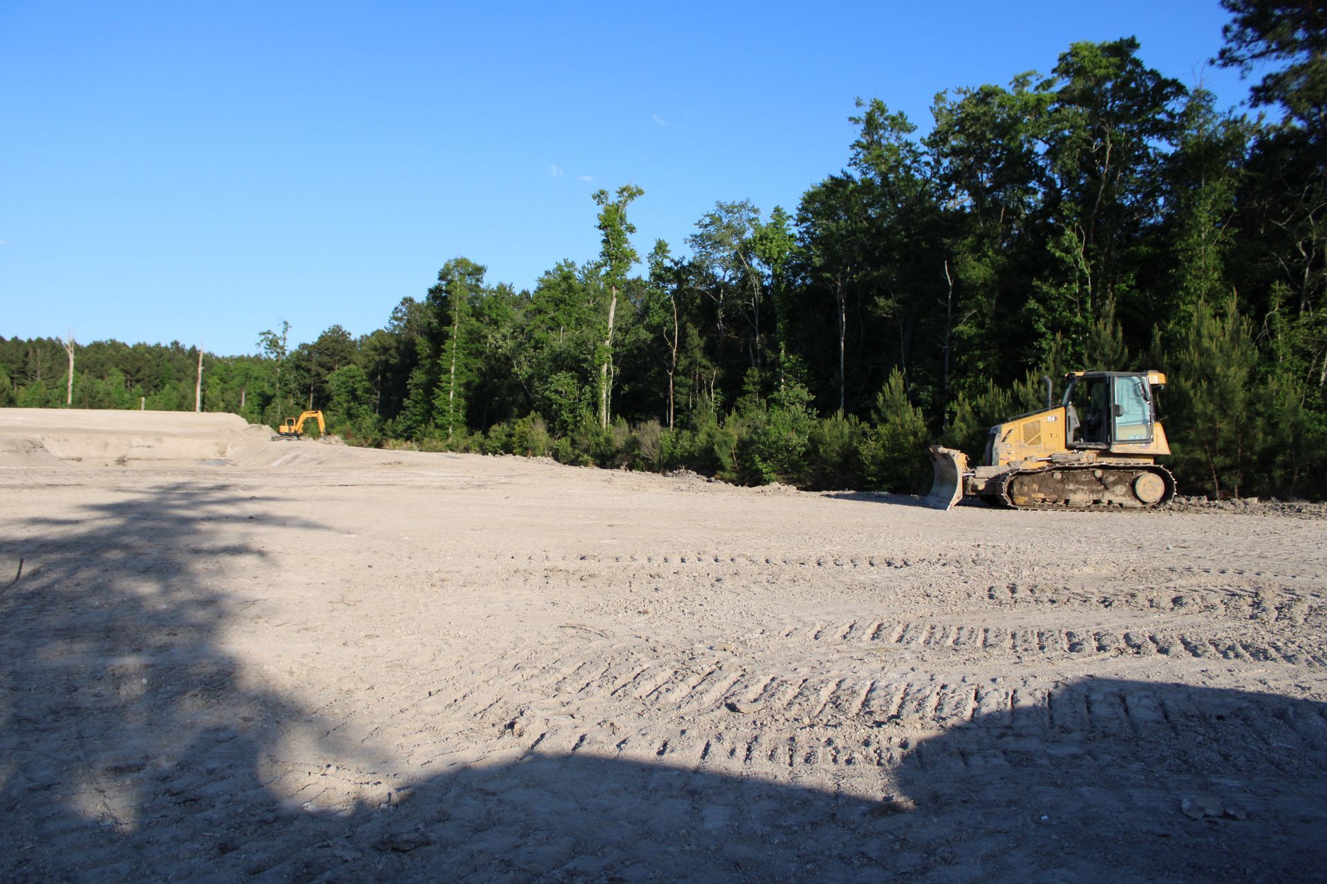 Bulldozer leveling a sandy construction site beside a tree line under a clear blue sky