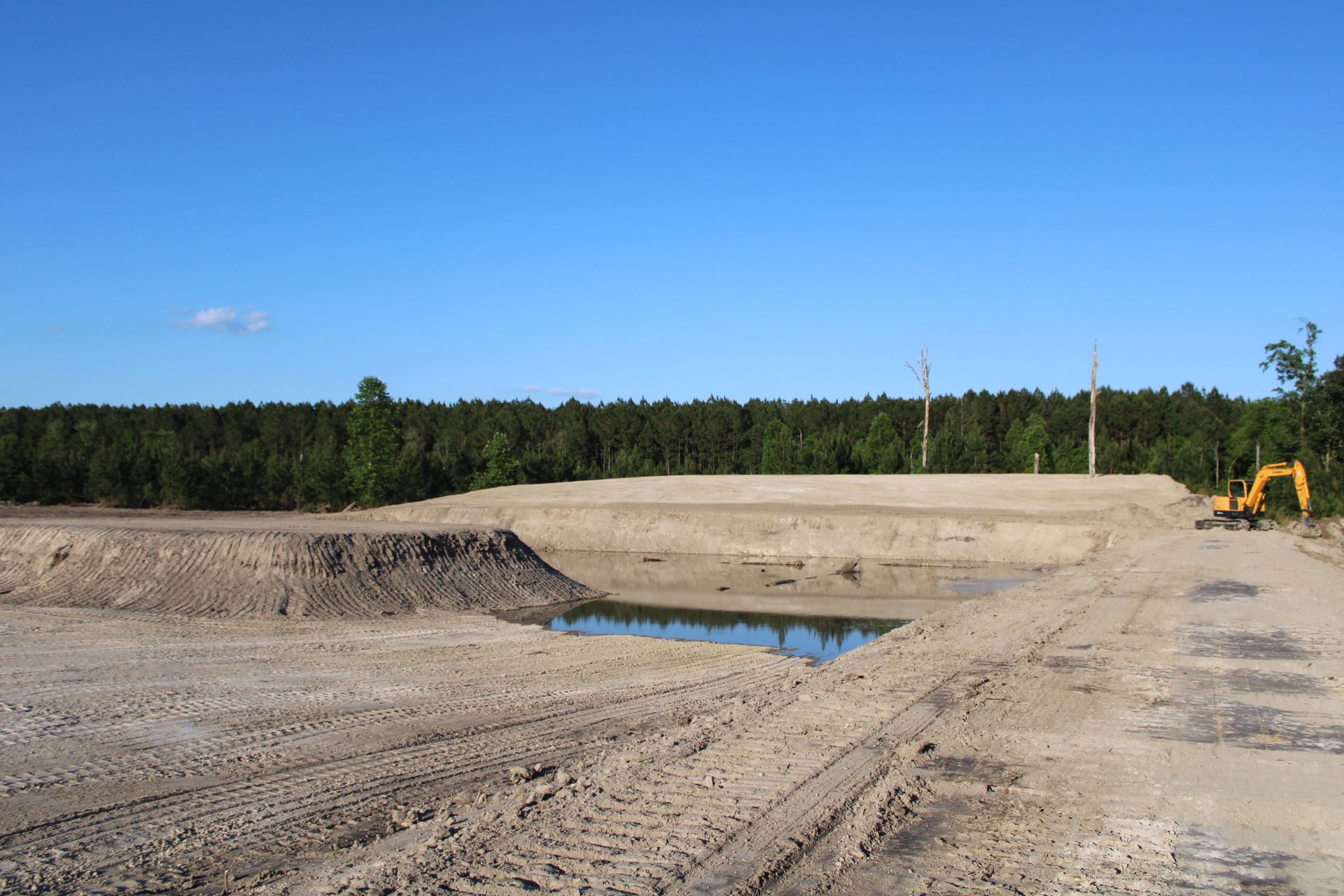 Excavation site with gravel piles, a shallow water pool, and an excavator under a clear blue sky.